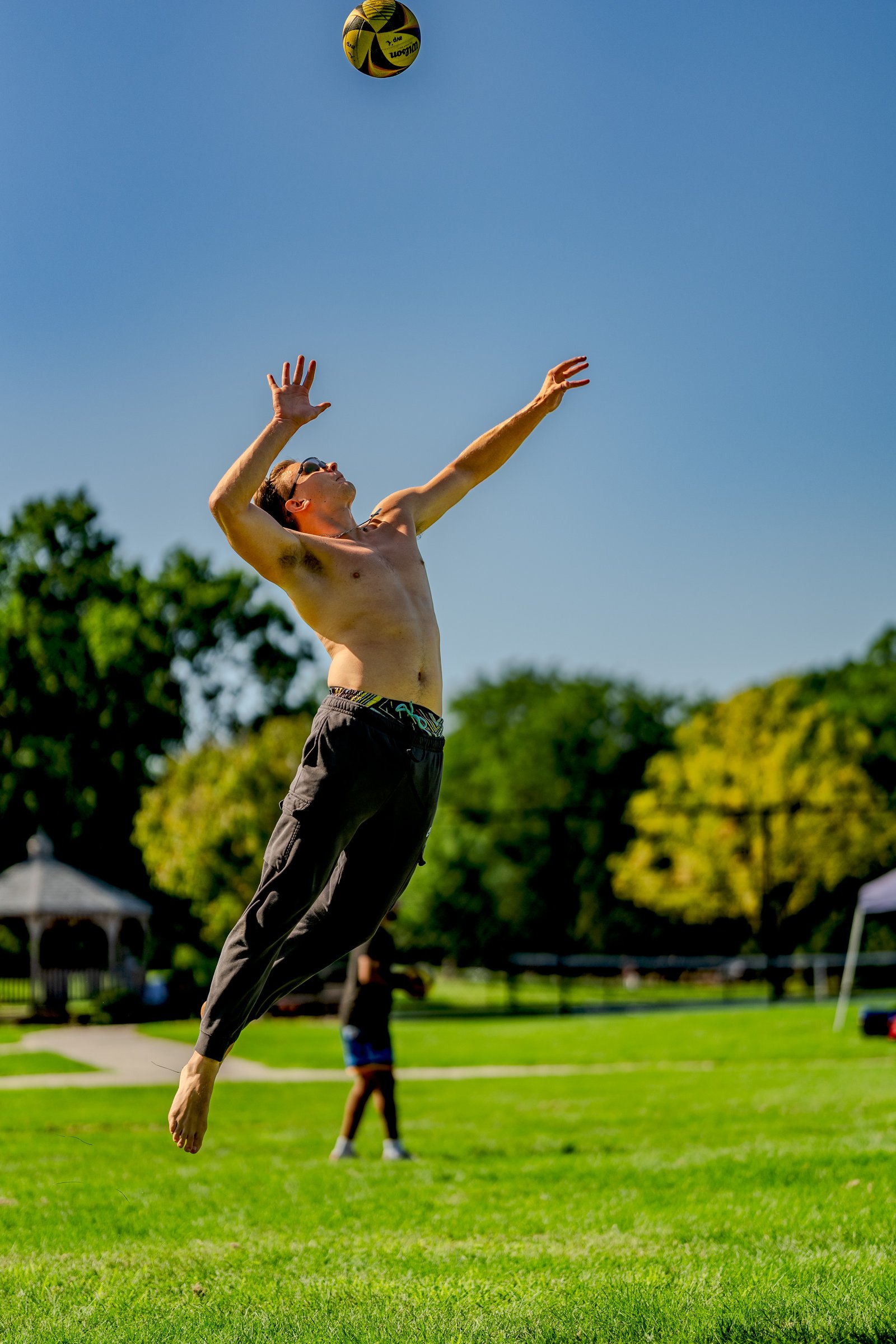 A player in black pants jumps to hit a volleyball against a clear blue sky with trees in the background.