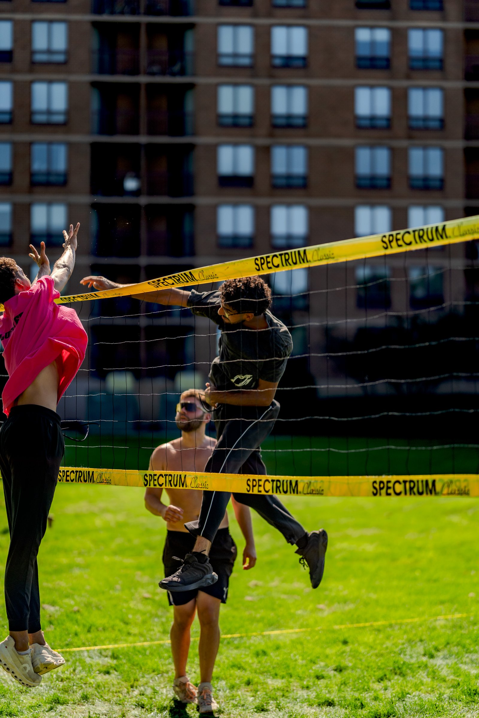 A player in a pink top jumps to hit a volleyball, with another player in a dark shirt reaching over the net on a sunny day.