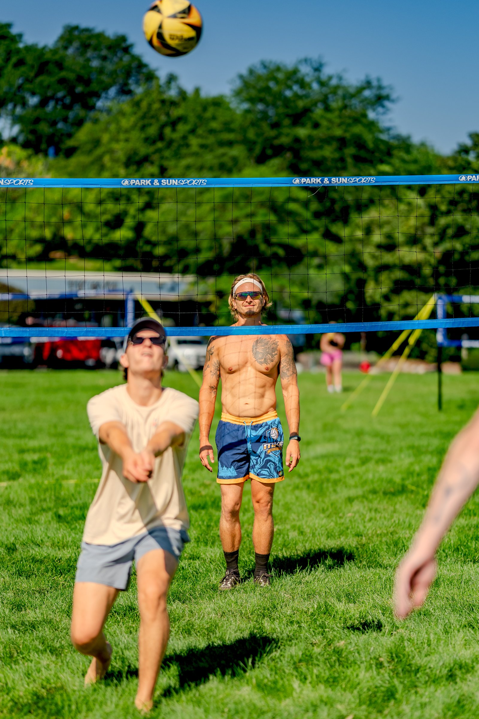 A player in a tan shirt and gray shorts passes a volleyball as a player in blue shorts stands by the net.