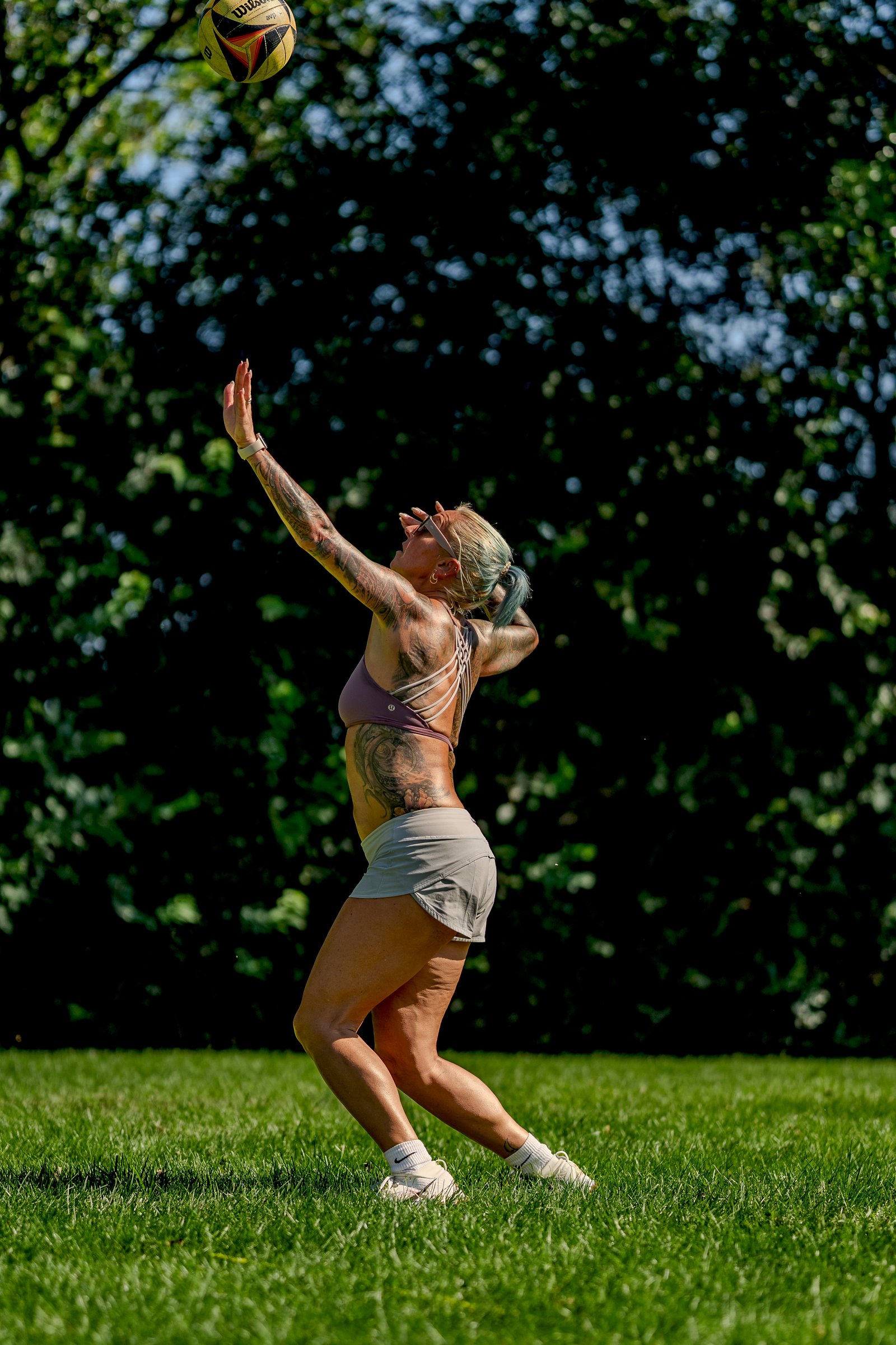 A player in a purple top and grey shorts reaches for a volleyball against a dark, leafy background.