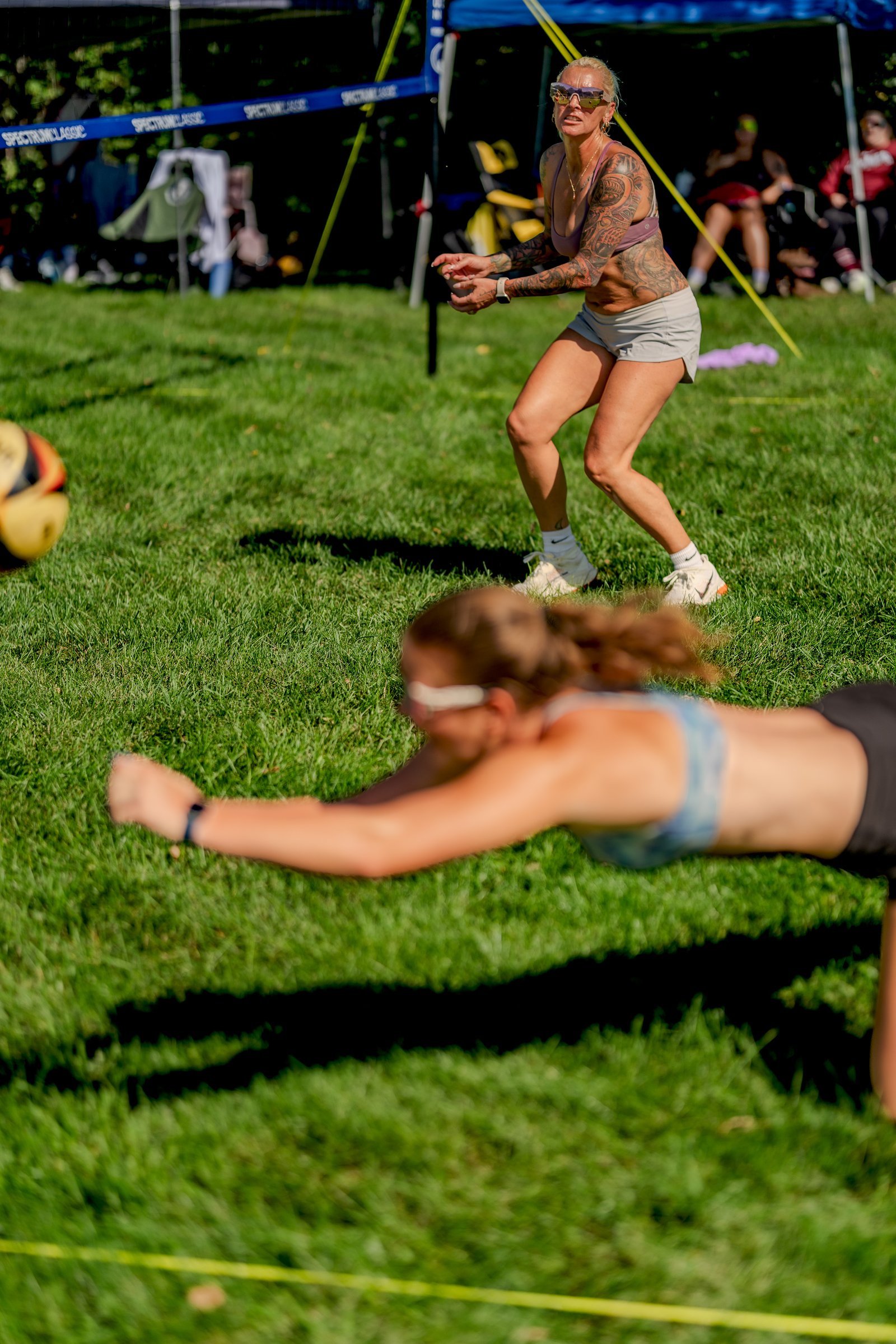 A player in a blue top dives for a volleyball while another player in a purple top watches.