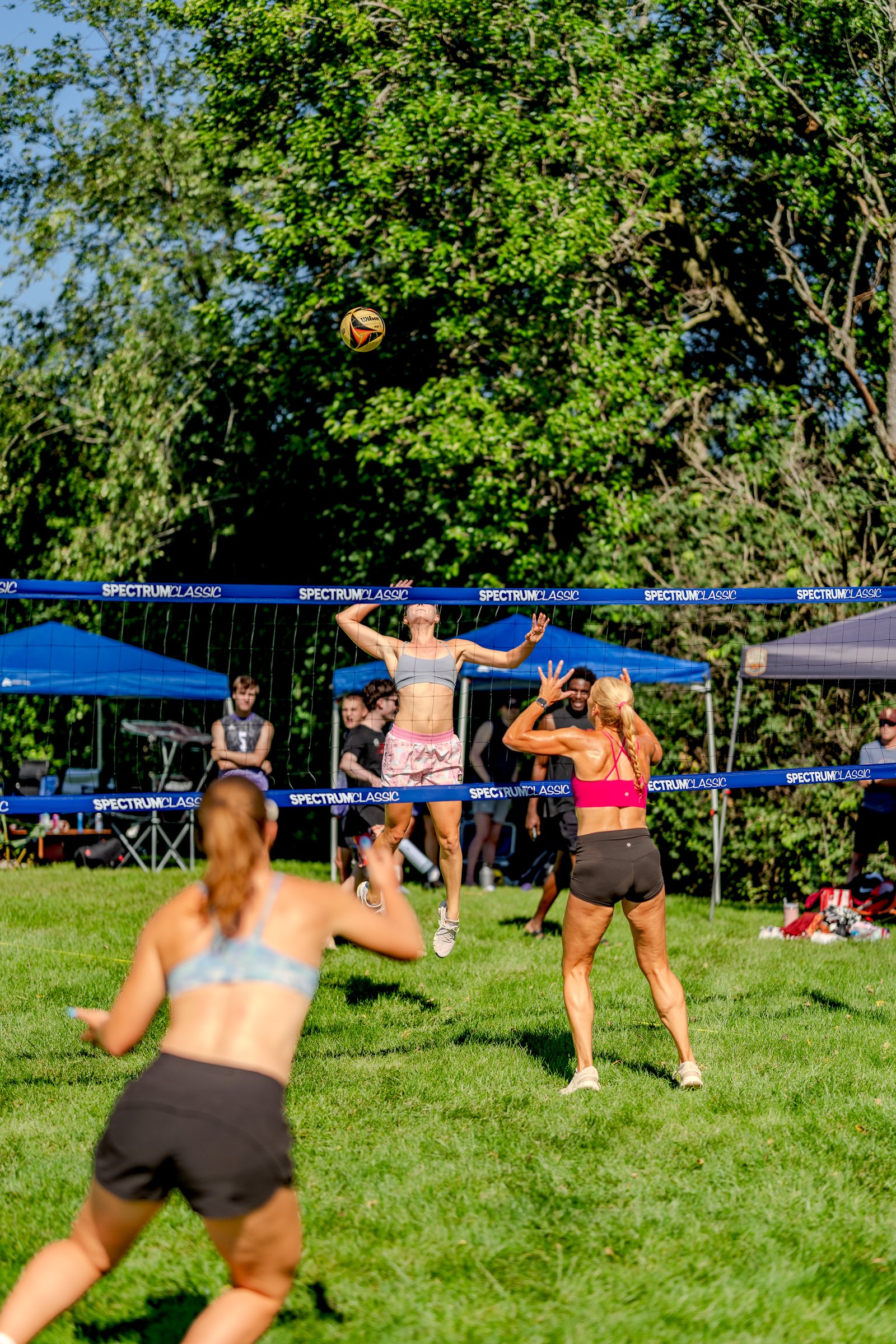 A player in a grey top jumps to hit the ball over the net while a player in a pink top prepares to receive, with other players visible.