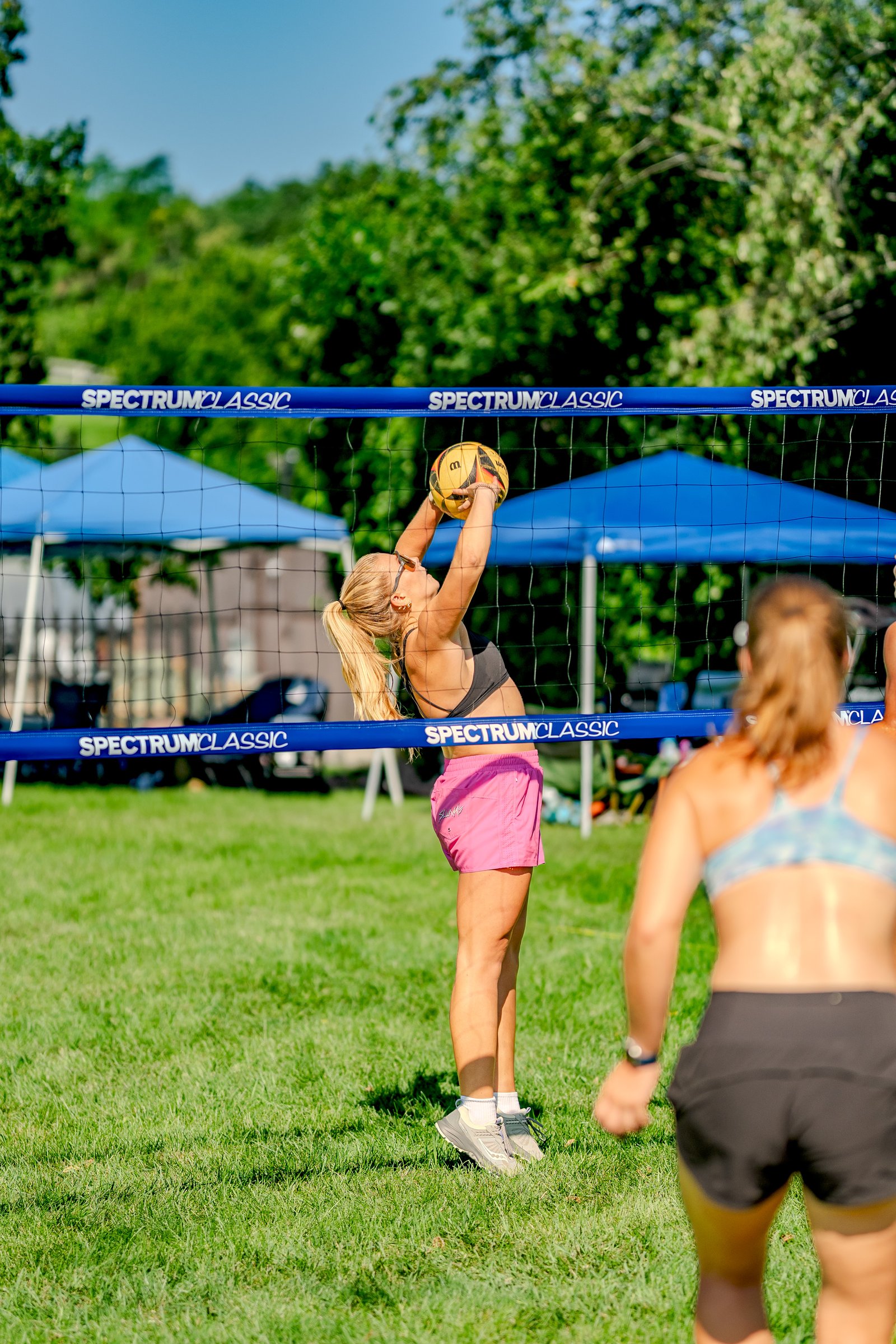A player in pink shorts and a black top sets the ball over the net during a volleyball game, with another player in a blue and white top visible.