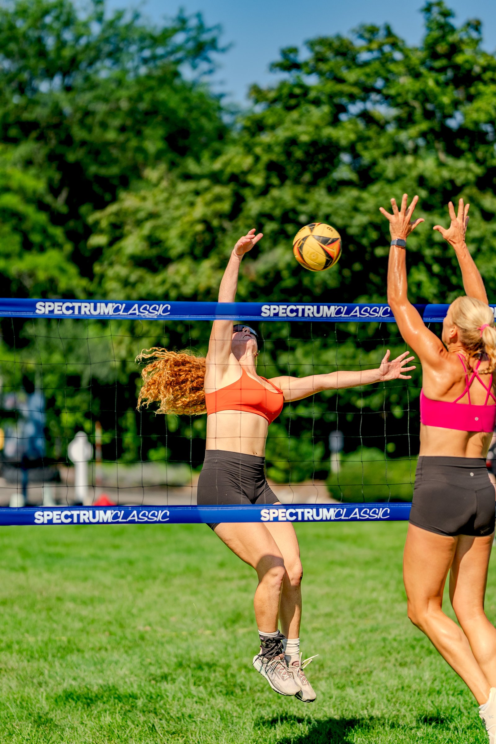 A player in an orange top jumps to hit a volleyball over the net, with a player in a pink top reaching up to block.