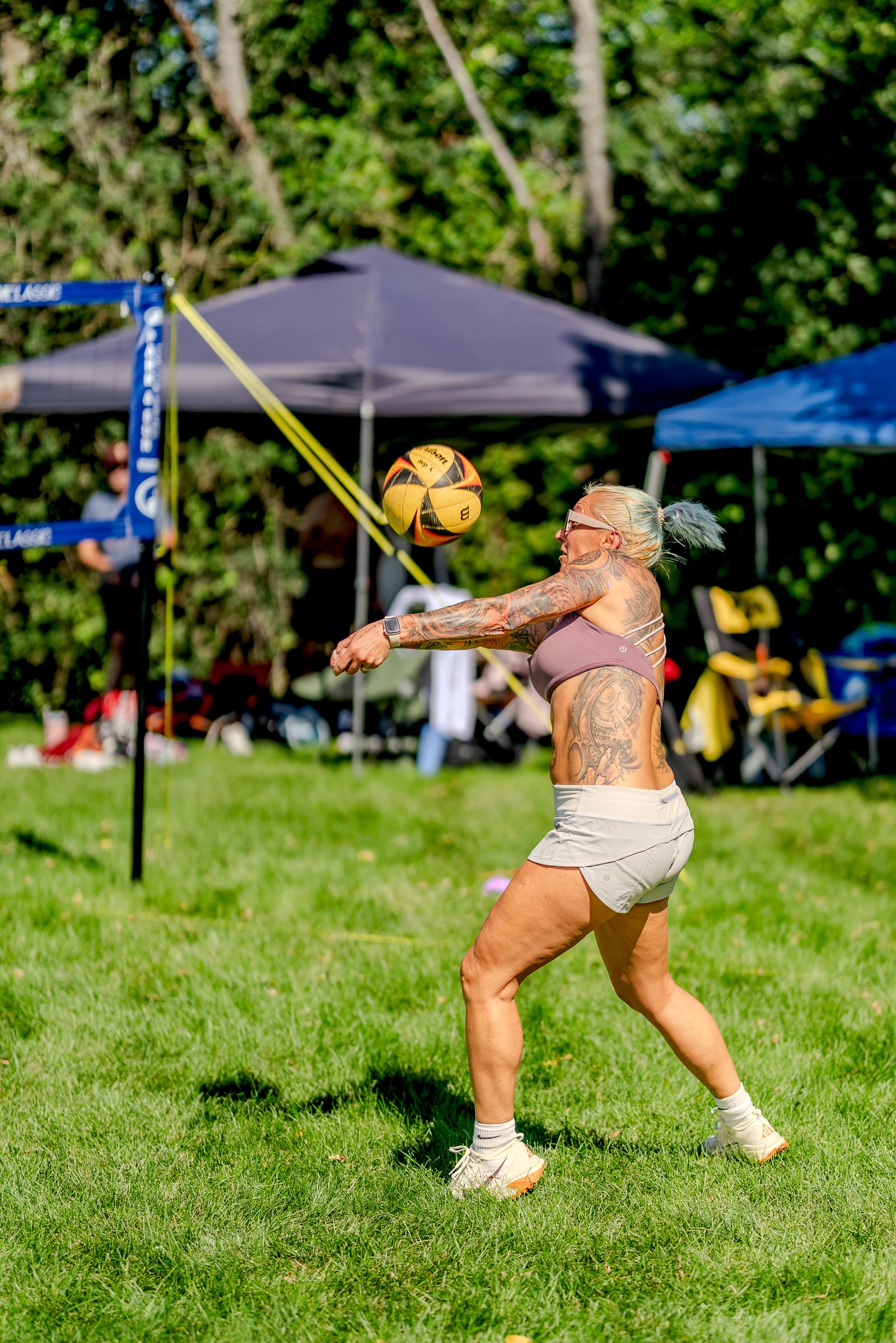 A player in a mauve top and white shorts serves a volleyball during a tournament on a grassy field.
