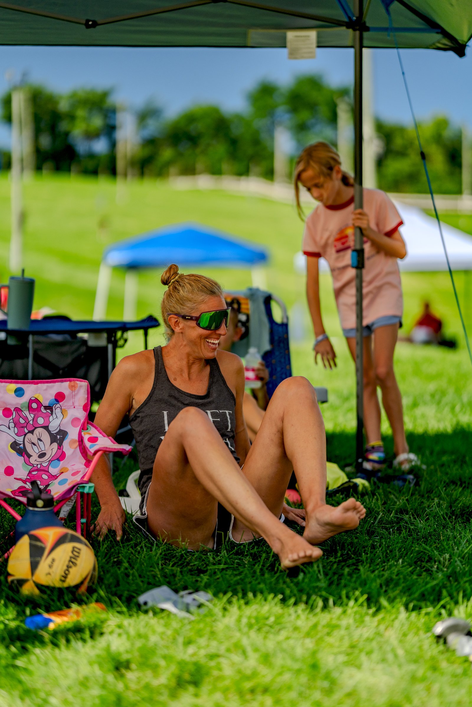 A player in a dark gray tank top sits on the grass next to a Minnie Mouse chair, while a player in a pink shirt stands nearby.