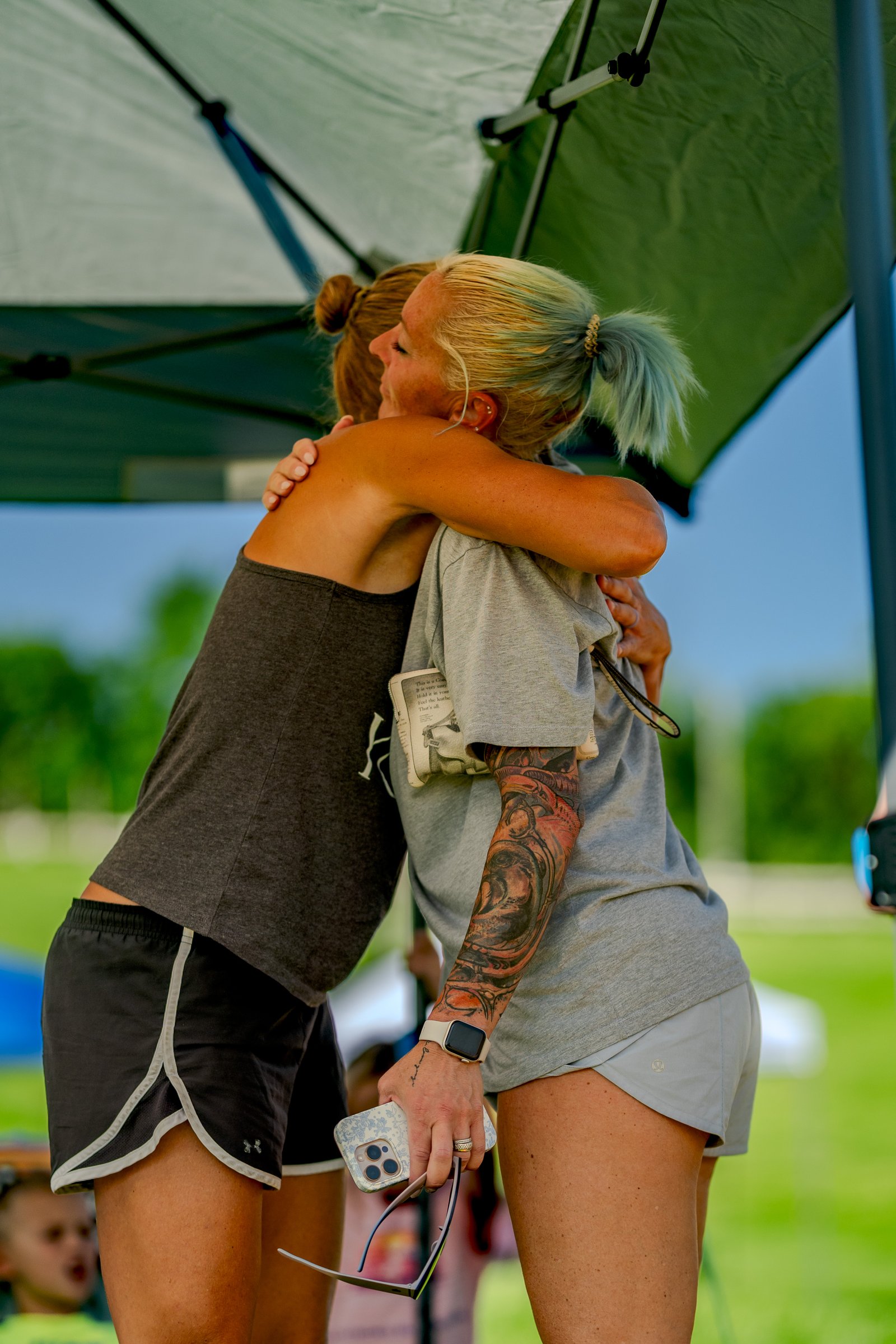 Two players hug under a tent. One player wears a dark grey tank top and black shorts, the other a grey t-shirt and light grey shorts.