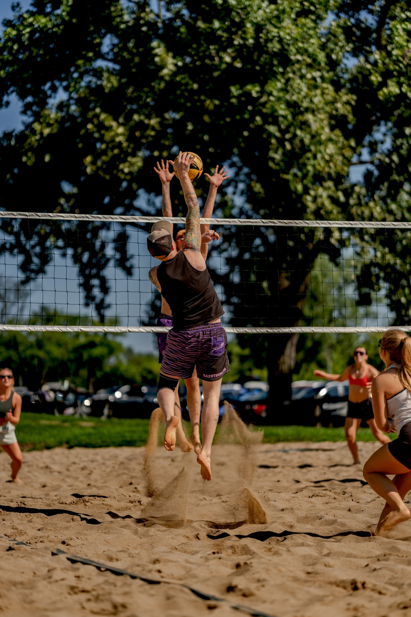 Players in black and purple jump at the net during a volleyball game, with sand flying.