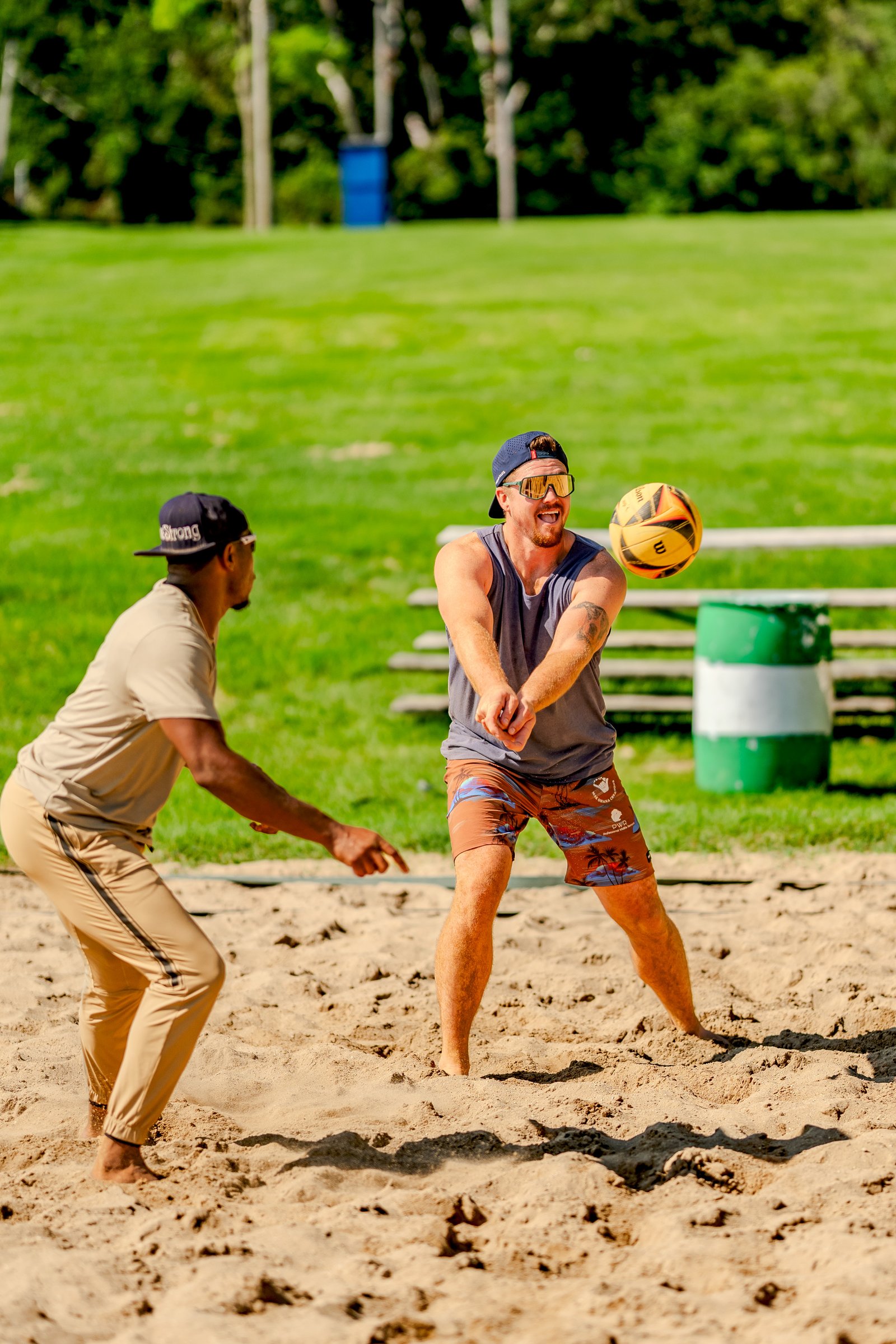 A player in a gray tank top and brown shorts prepares to pass a volleyball on a sand court, with another player in a tan shirt watching.