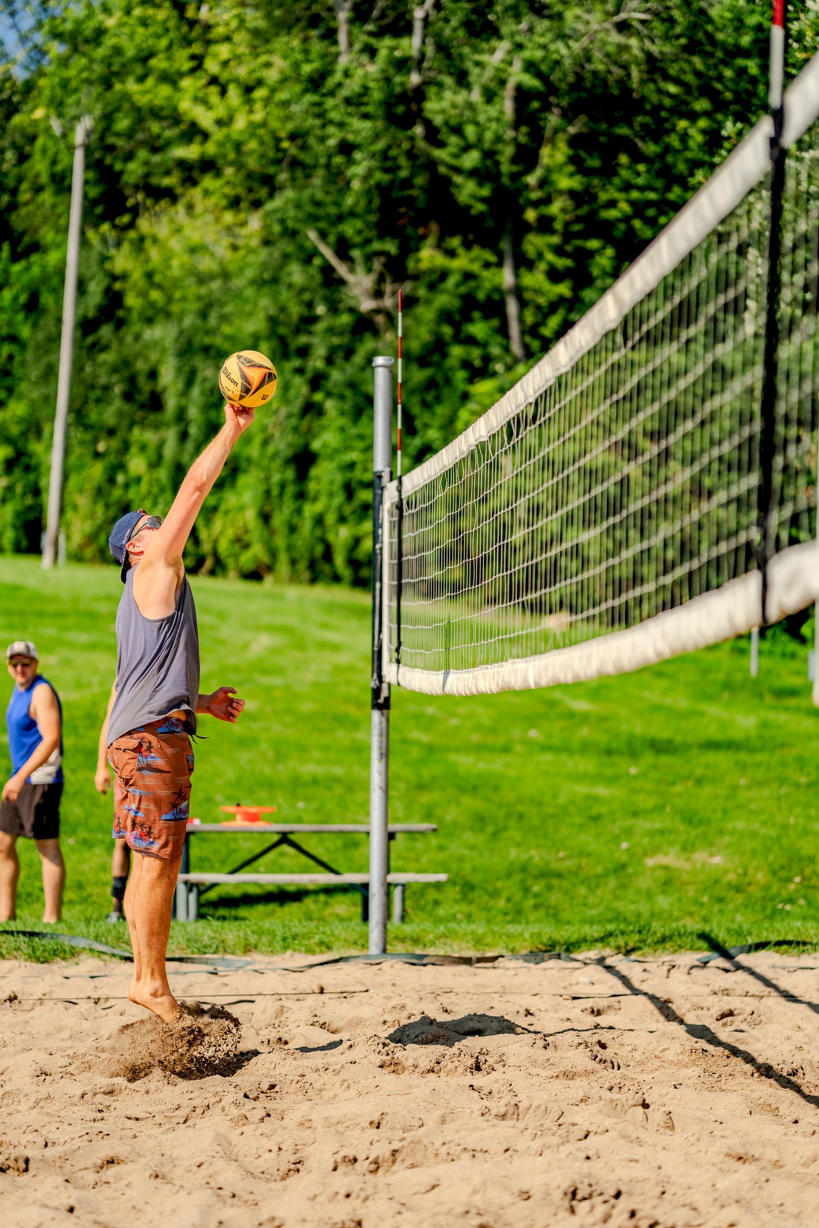 A player in a gray tank top and patterned shorts serves a volleyball on a sand court with a net and trees in the background.