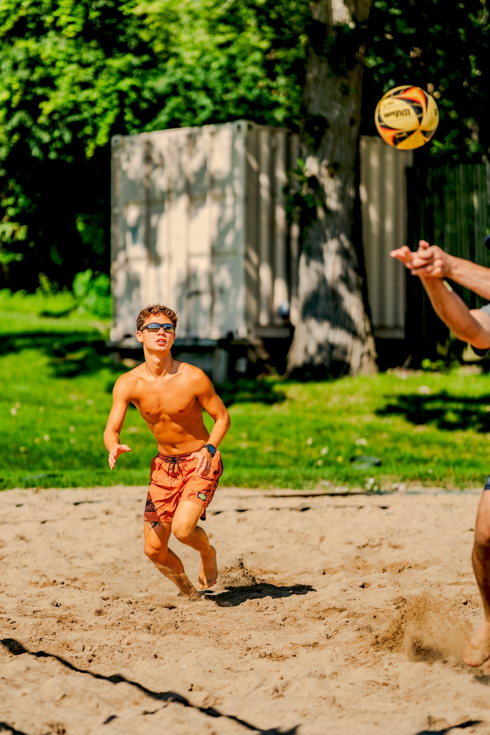 A player in orange shorts moves on the sand as a volleyball flies overhead near a tree and shipping containers.