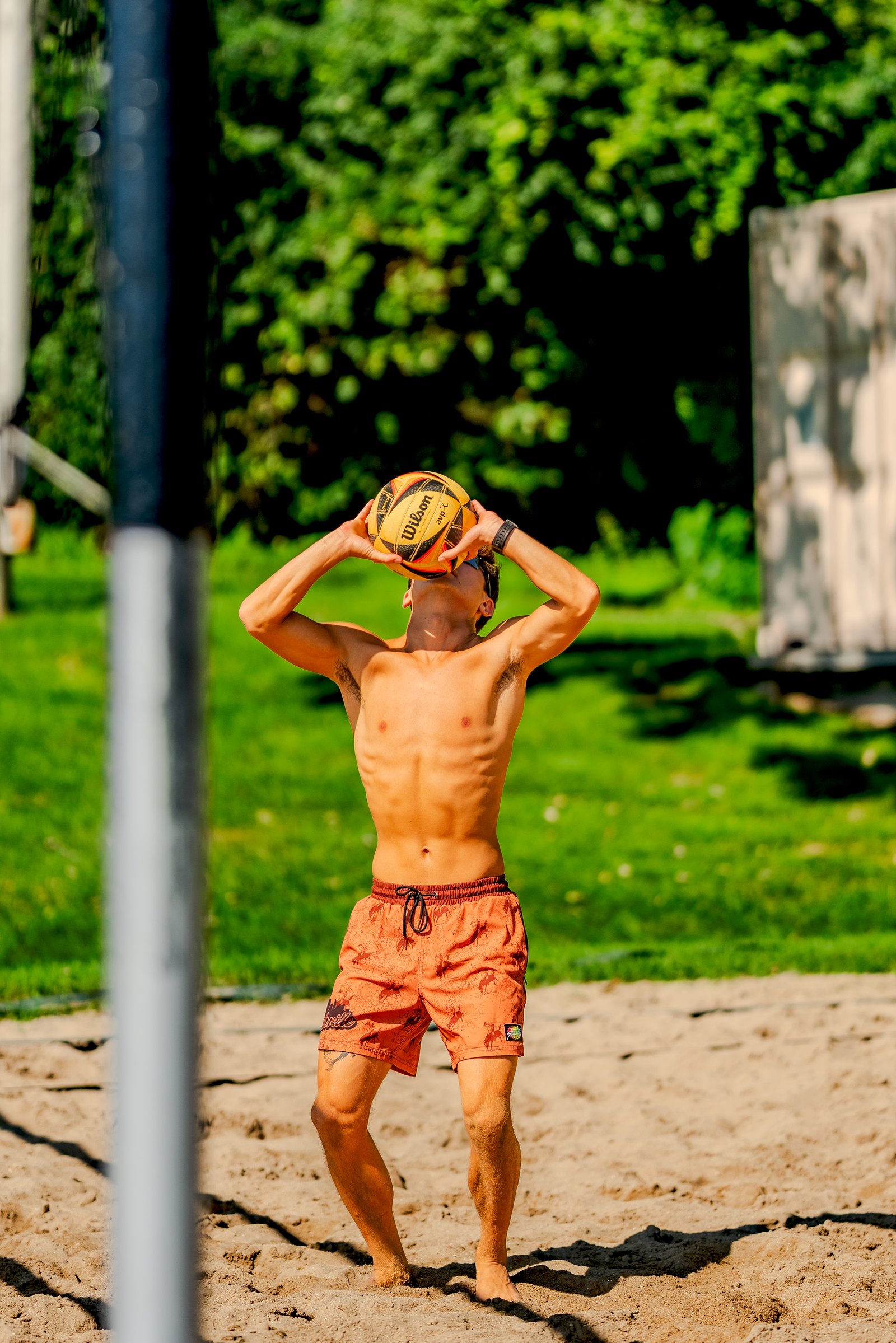 A player in orange shorts prepares to serve a volleyball on a sandy court with green trees in the background.