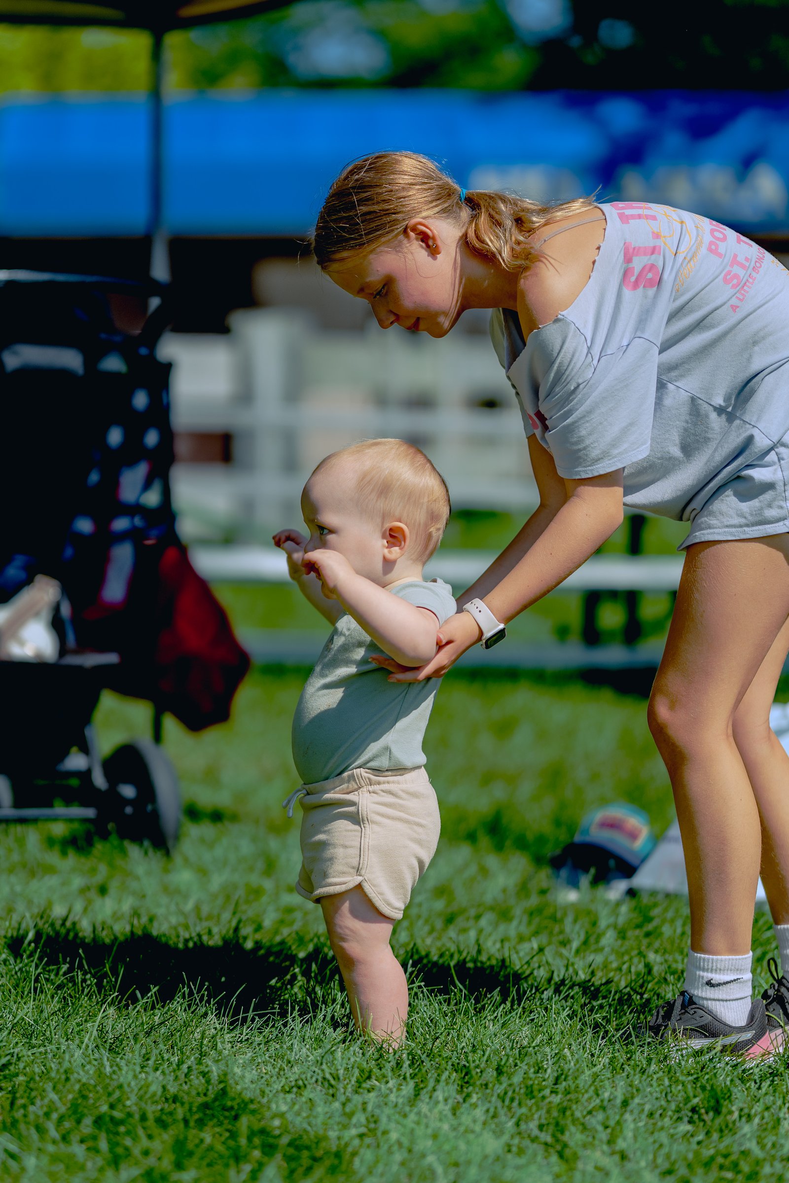 A player in a light blue shirt and shorts bends down to assist a baby standing on the grass.
