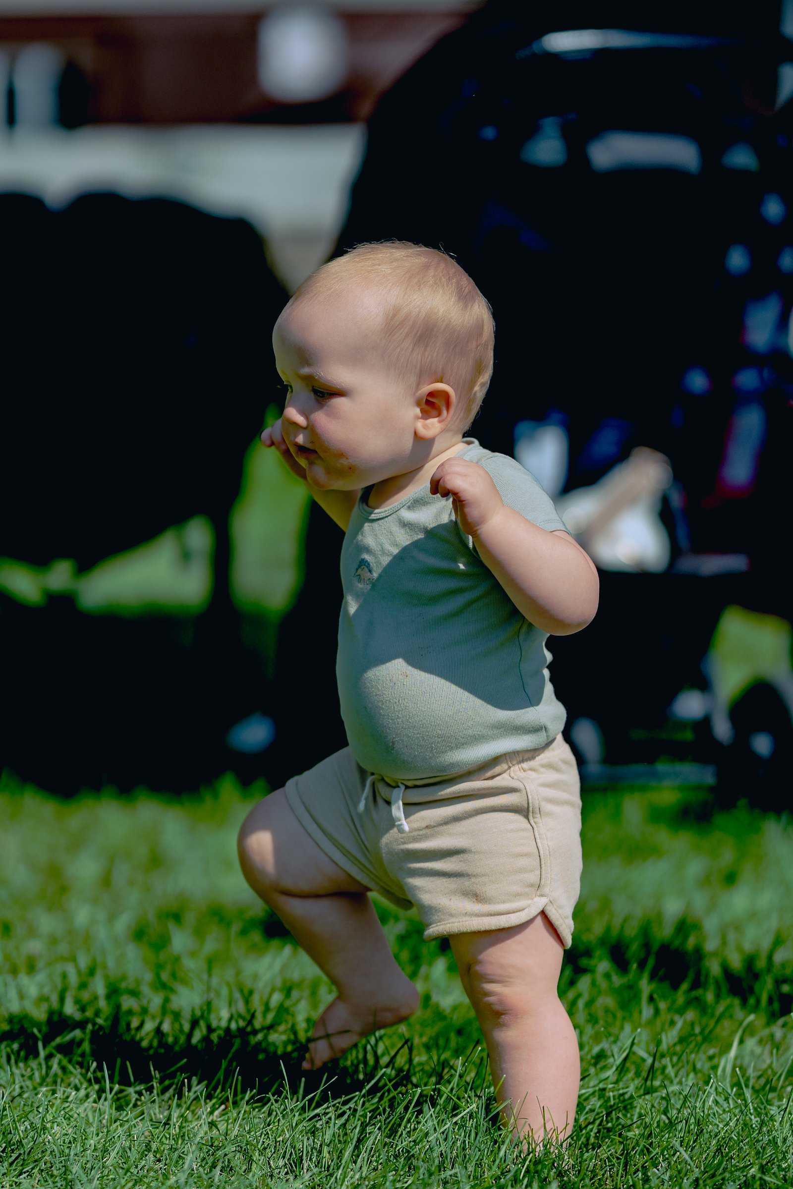 A baby in a light green shirt and tan shorts walks on grass with blurred figures in the background.