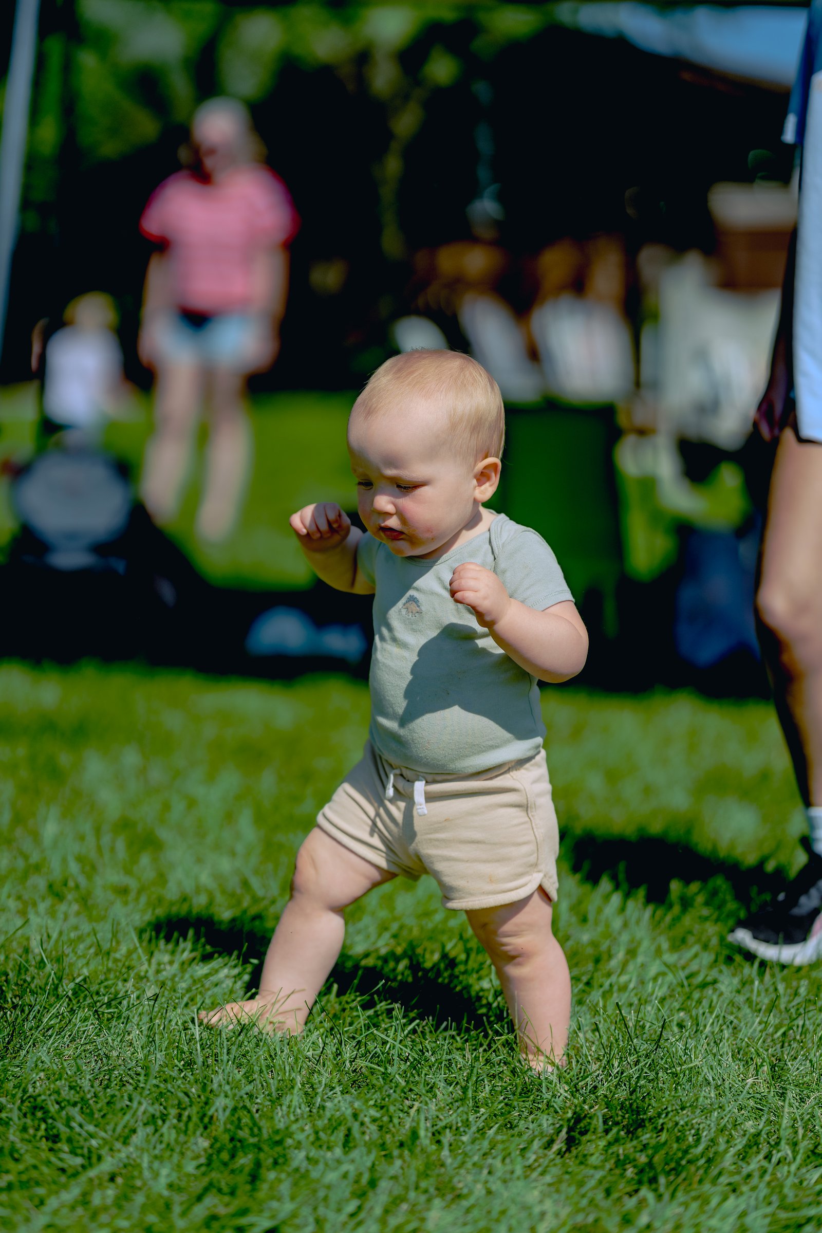 A baby in a light green shirt and tan shorts stands on grass with fists raised, while blurred figures are visible in the background.
