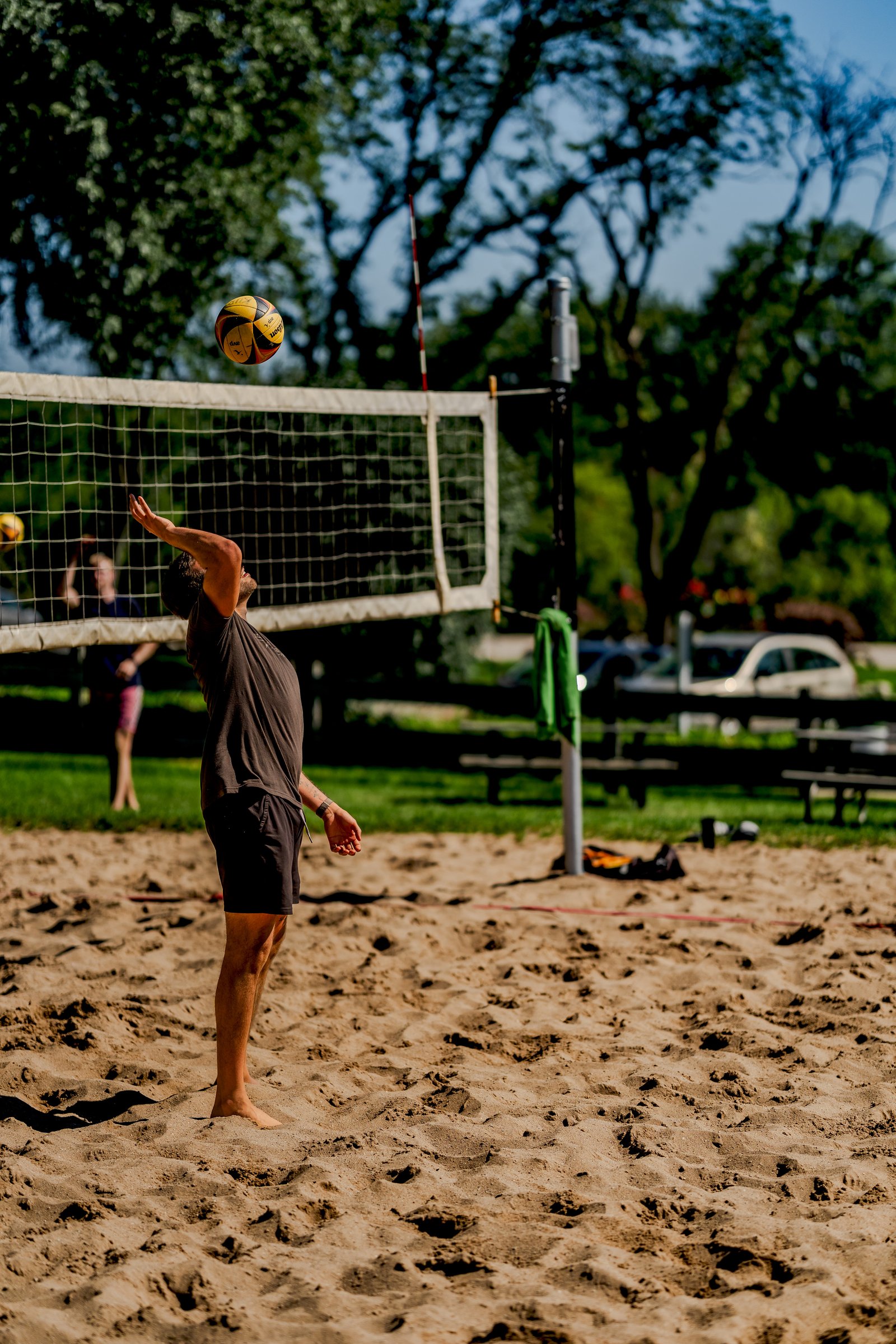 A player in a dark shirt and black shorts serves a volleyball on a sand court with a net and trees in the background.