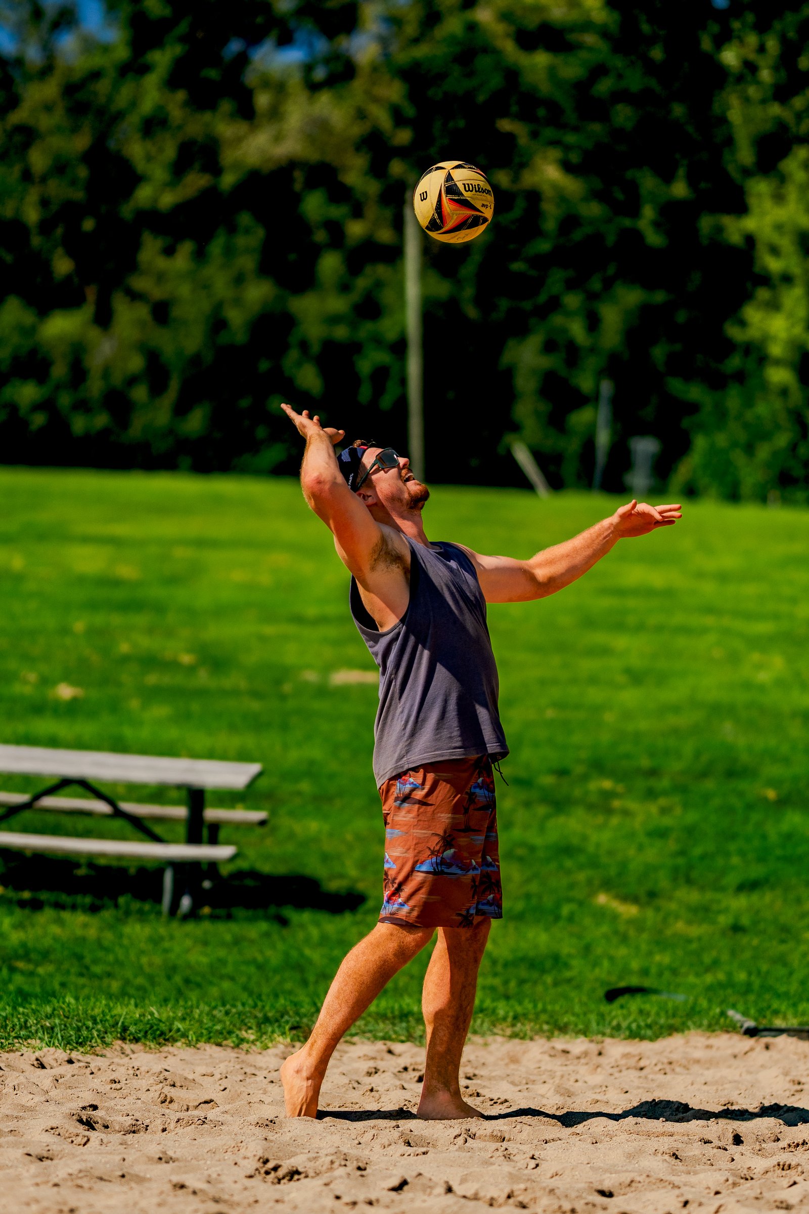 A player in a gray tank top and brown patterned shorts prepares to serve a volleyball on a sandy court.