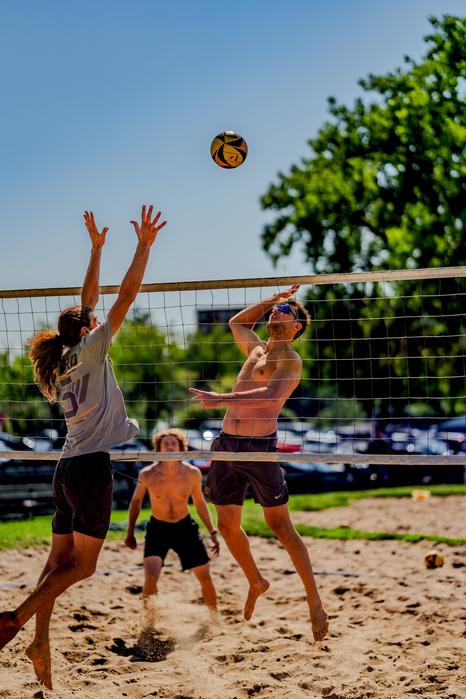 Players in grey and black shorts jump near the net as a volleyball is in the air during a tournament.