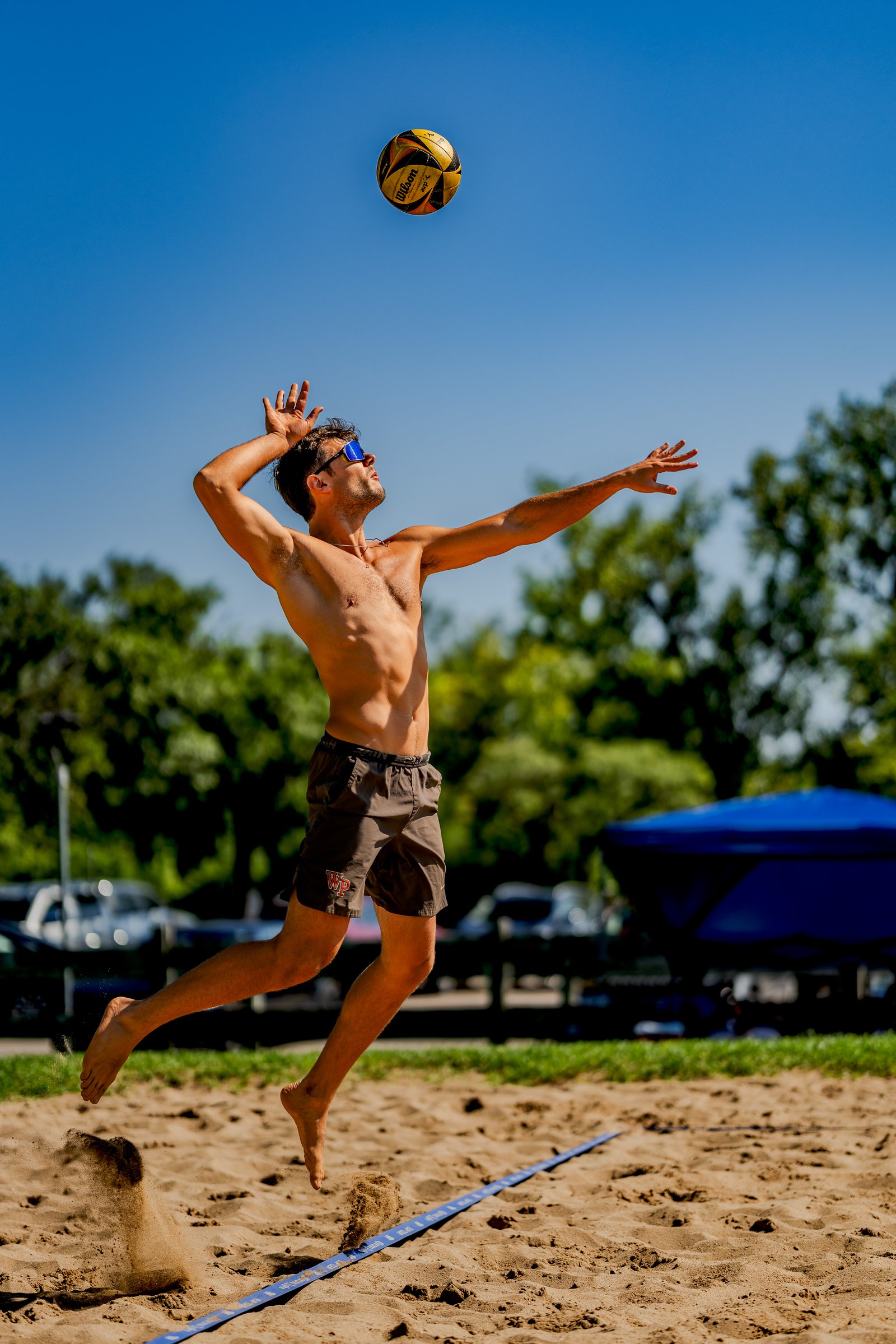A player in brown shorts jumps to hit a volleyball against a clear blue sky.