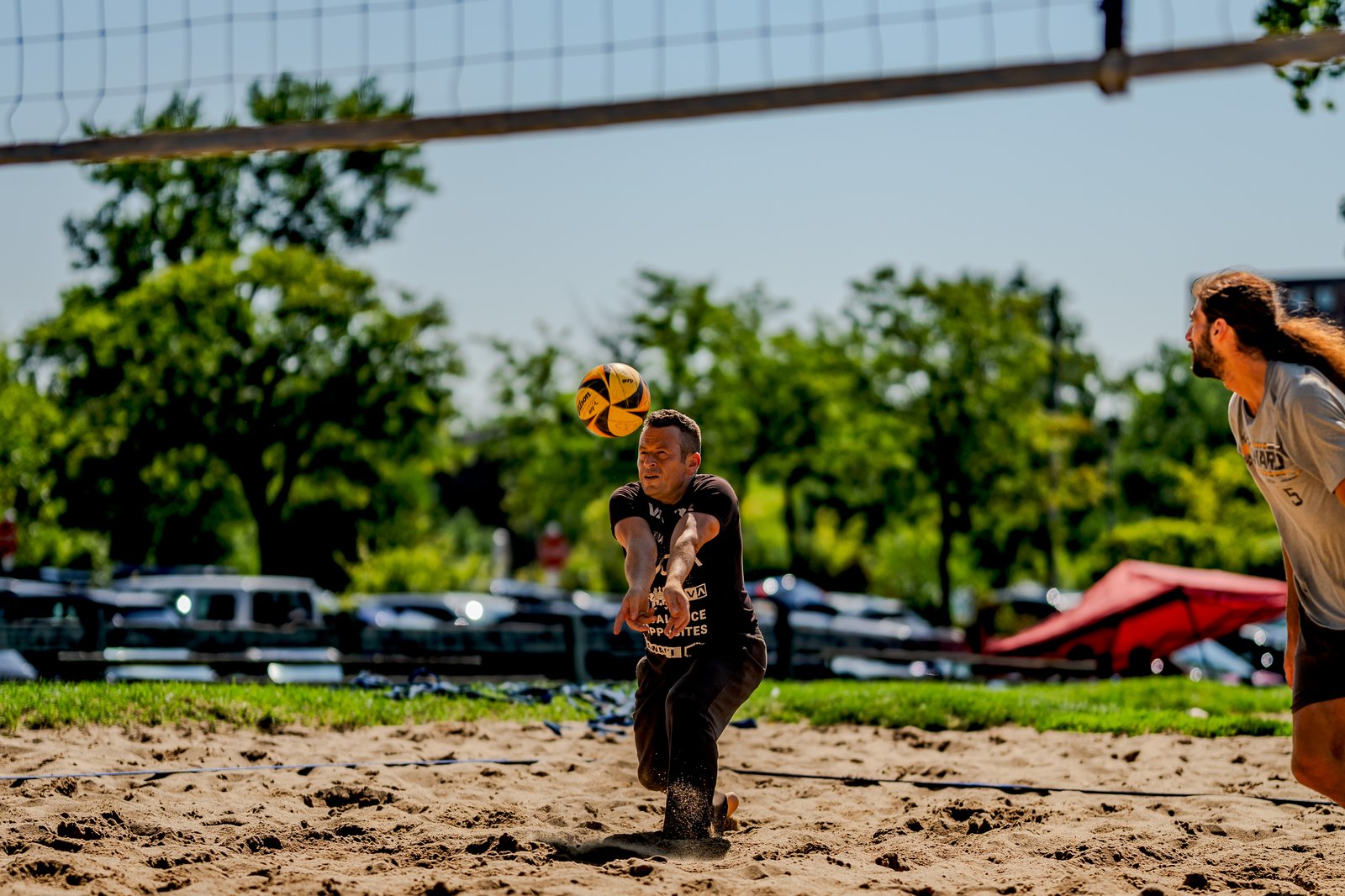 A player in black dives to pass a volleyball on a sand court, with another player in a gray shirt watching.