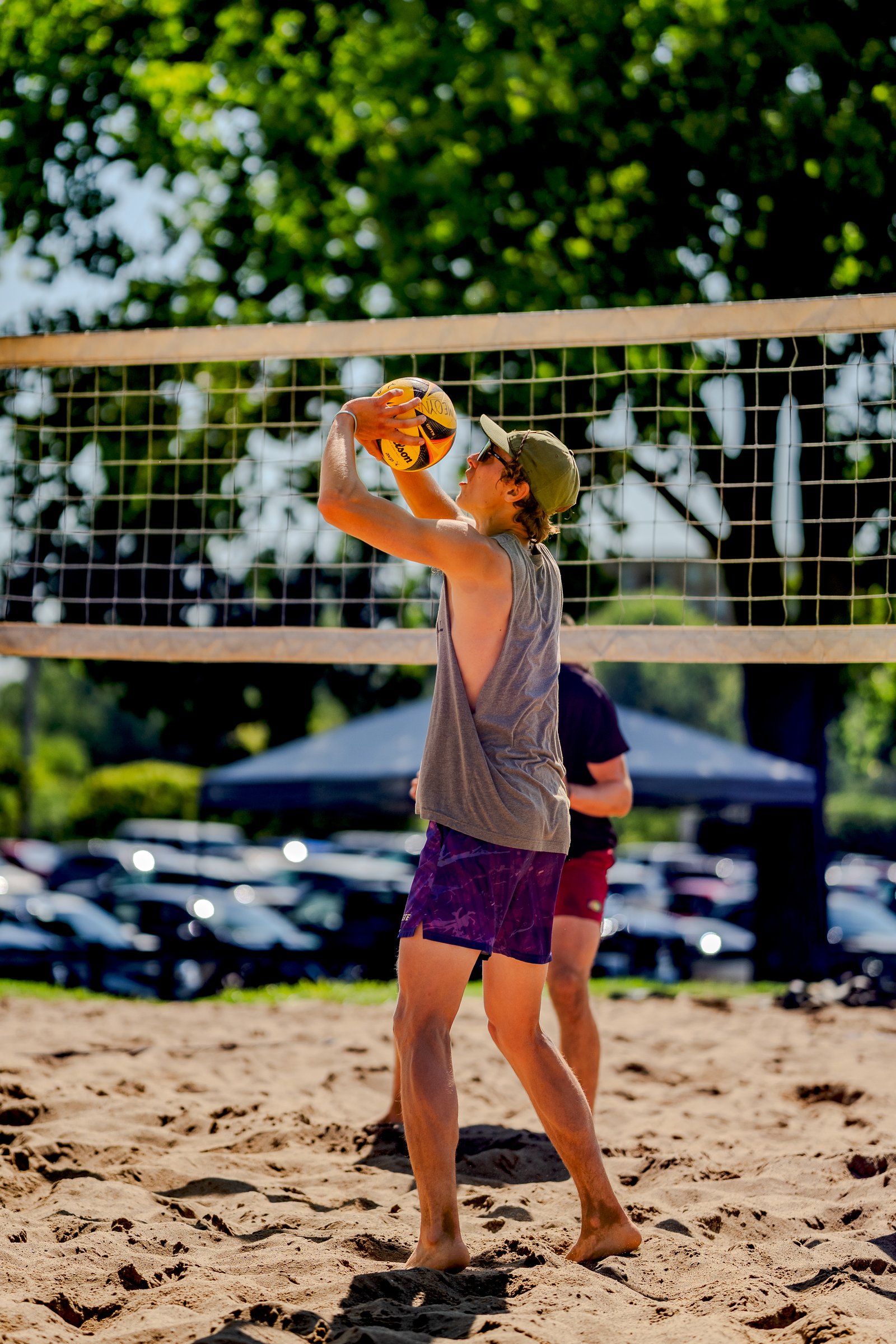 A player in a gray tank top and purple shorts prepares to set the volleyball on a sandy court.