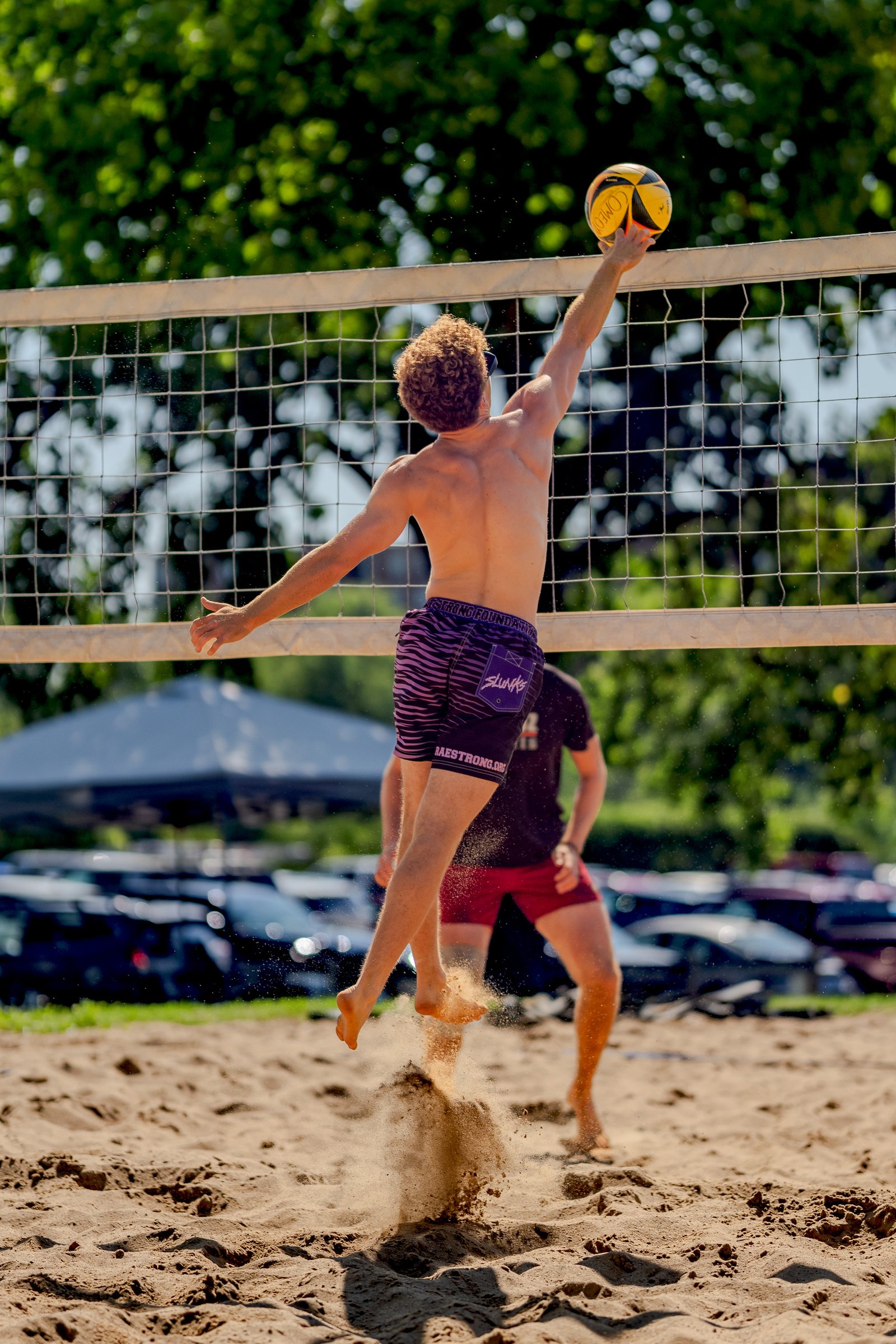 A player in purple striped shorts reaches for the volleyball over the net, with another player in red shorts behind him on the sand court.