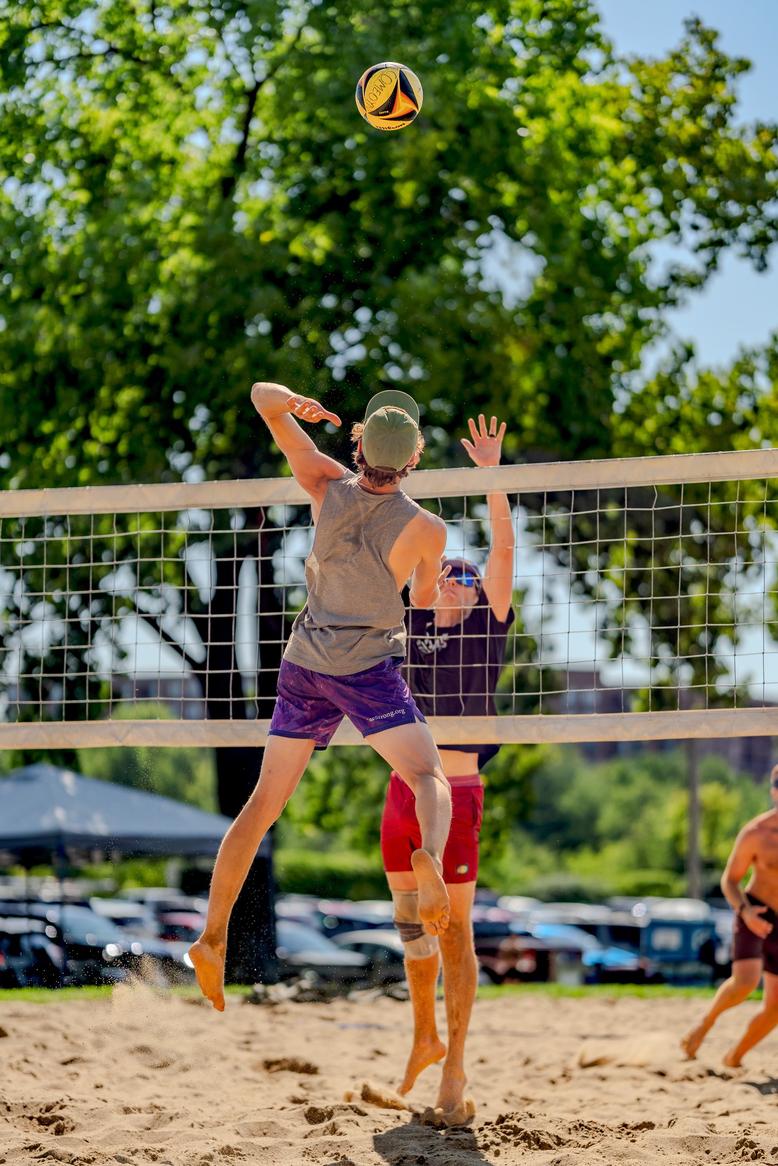 A player in a gray top and purple shorts jumps to hit a volleyball over the net, with a player in a black top and red shorts blocking.
