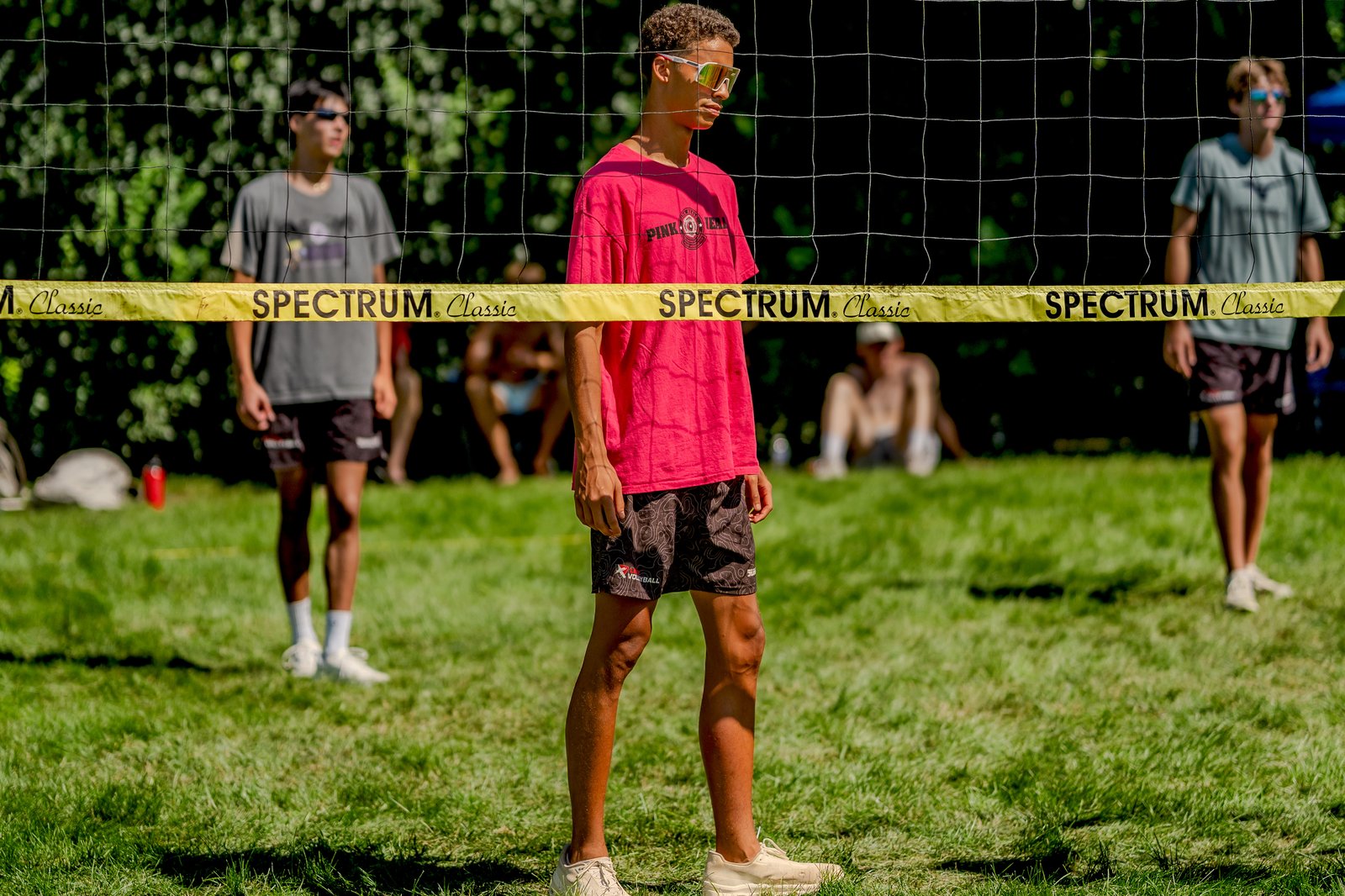 A player in a pink shirt and patterned shorts stands near the net, with other players in gray and blue shirts visible in the background.