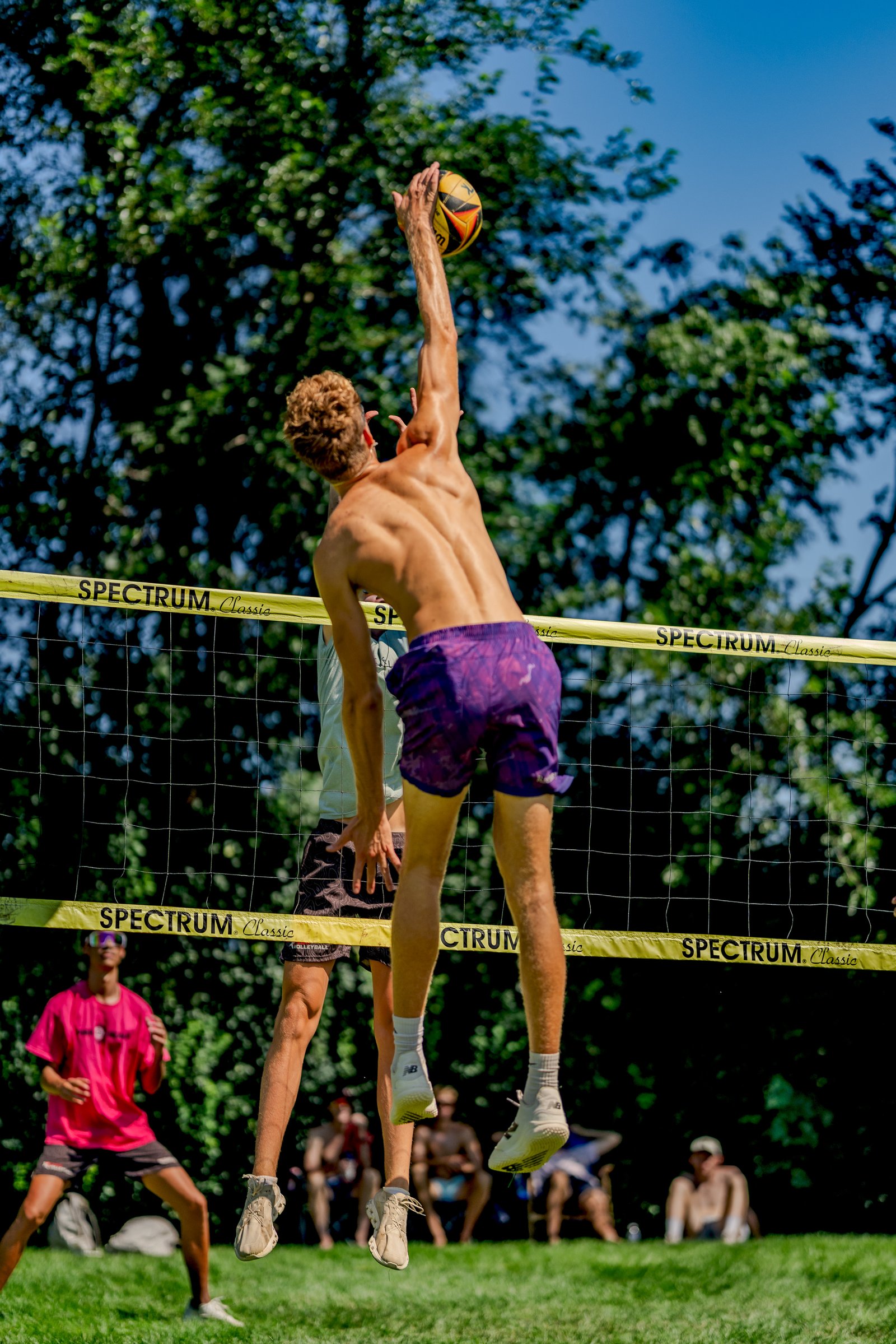 A player in purple shorts jumps to hit a volleyball over the net, with other players and spectators in the background.