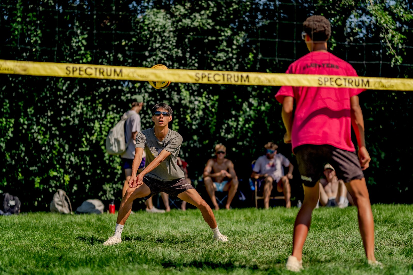 A player in a grey shirt prepares to dig a volleyball during a tournament, with other players watching from the sidelines.