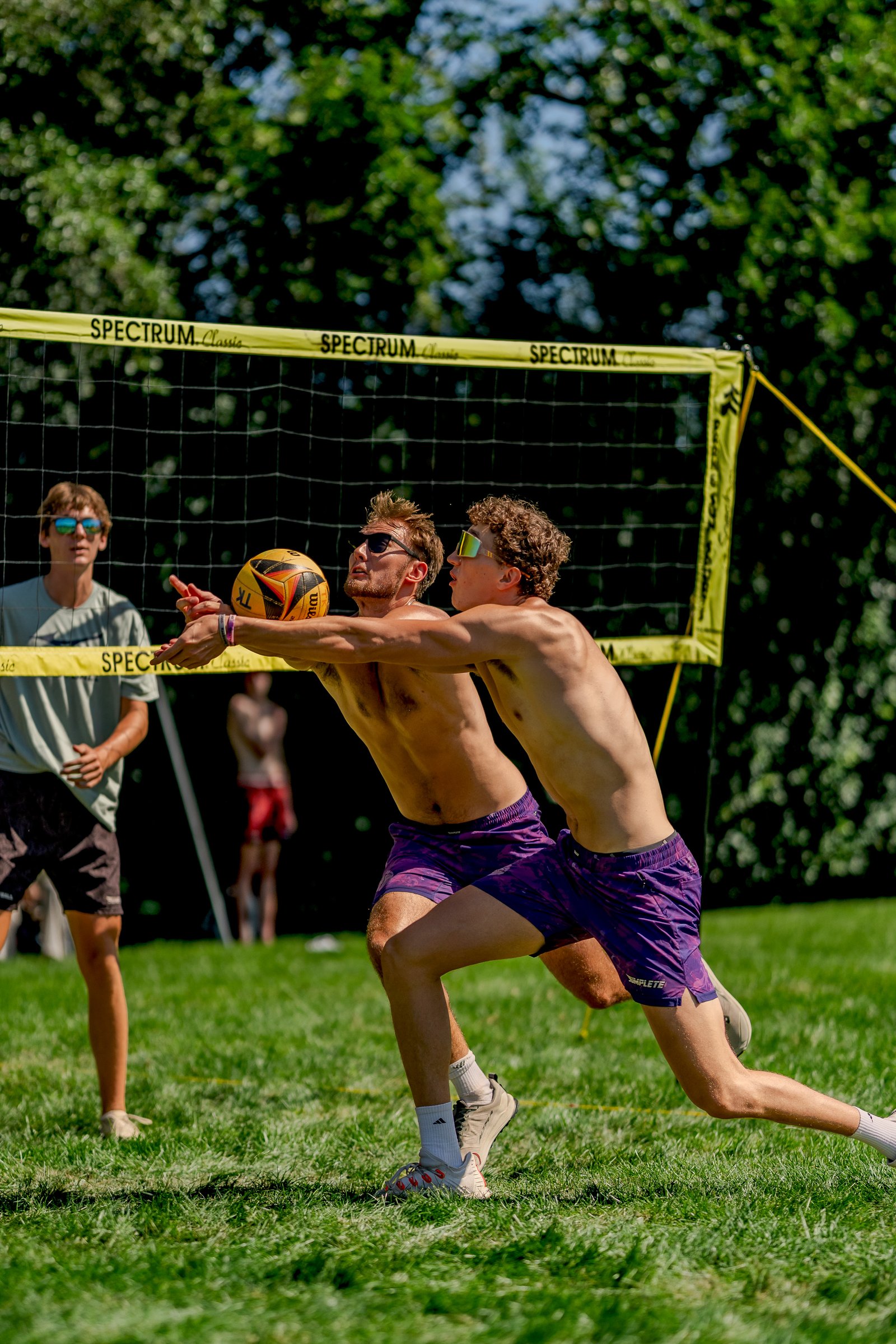 Two players in purple shorts and a player in a light green shirt play volleyball on a grass court with a yellow net.