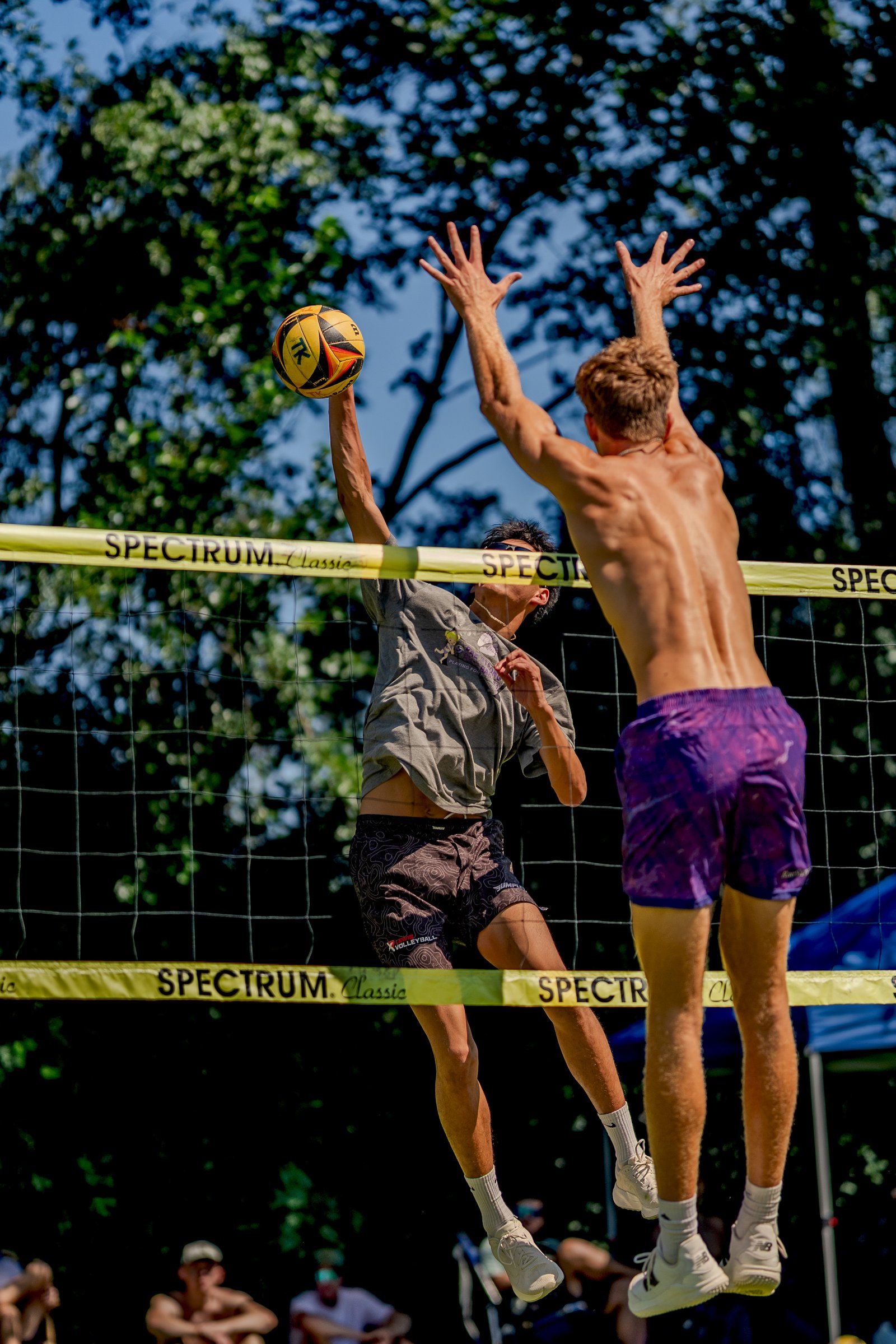 A player in a grey shirt and patterned shorts jumps to hit a volleyball, with another player in purple shorts reaching up.