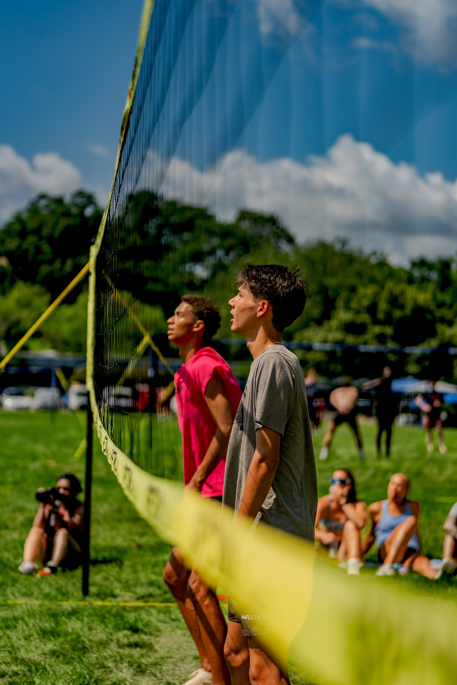 Two players in pink and gray shirts look up at the volleyball net on a sunny day with spectators in the background.