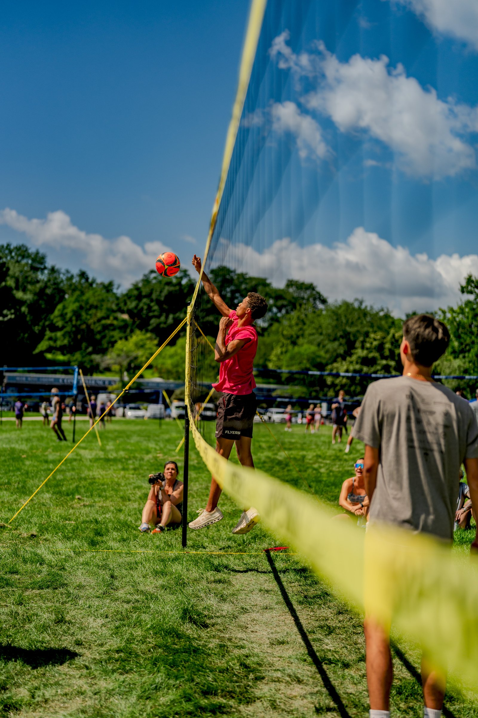 A player in a pink top and black shorts jumps to hit a volleyball over the net, with spectators watching on the grass.