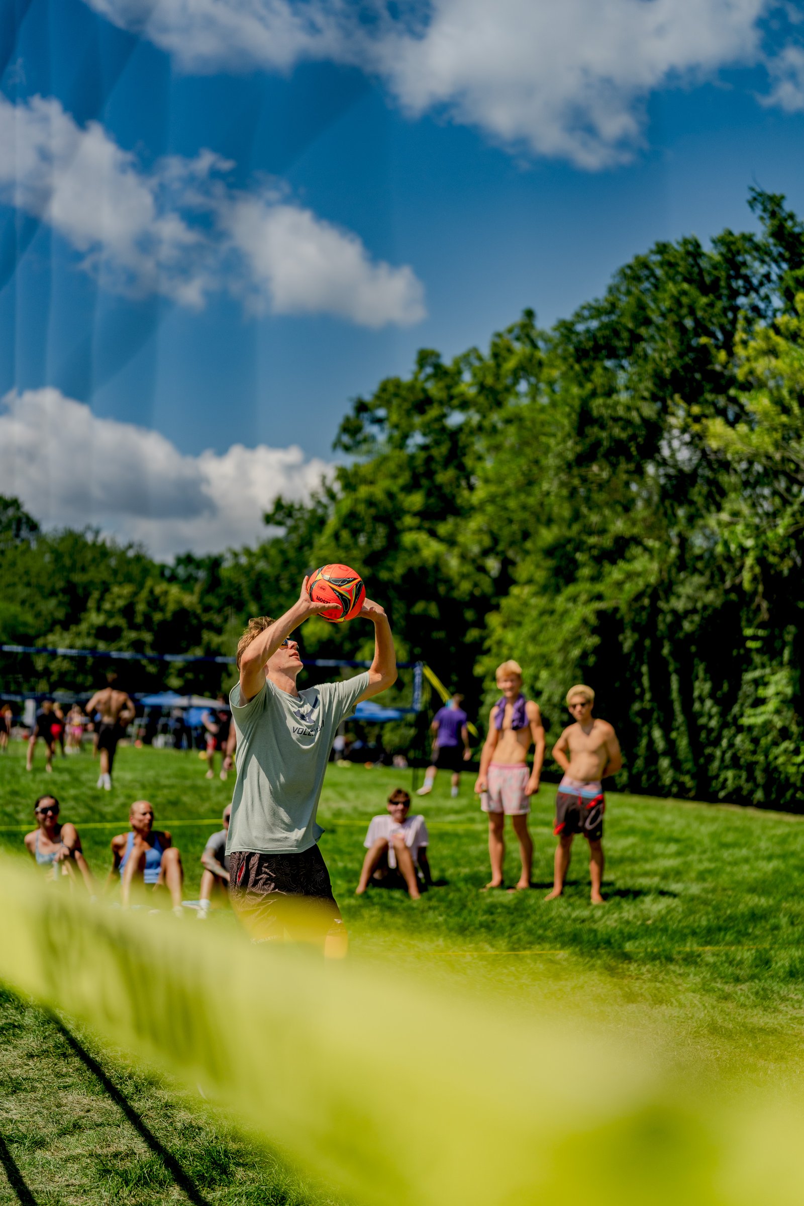 A player in a light green shirt prepares to serve a volleyball on a sunny day with spectators in the background.