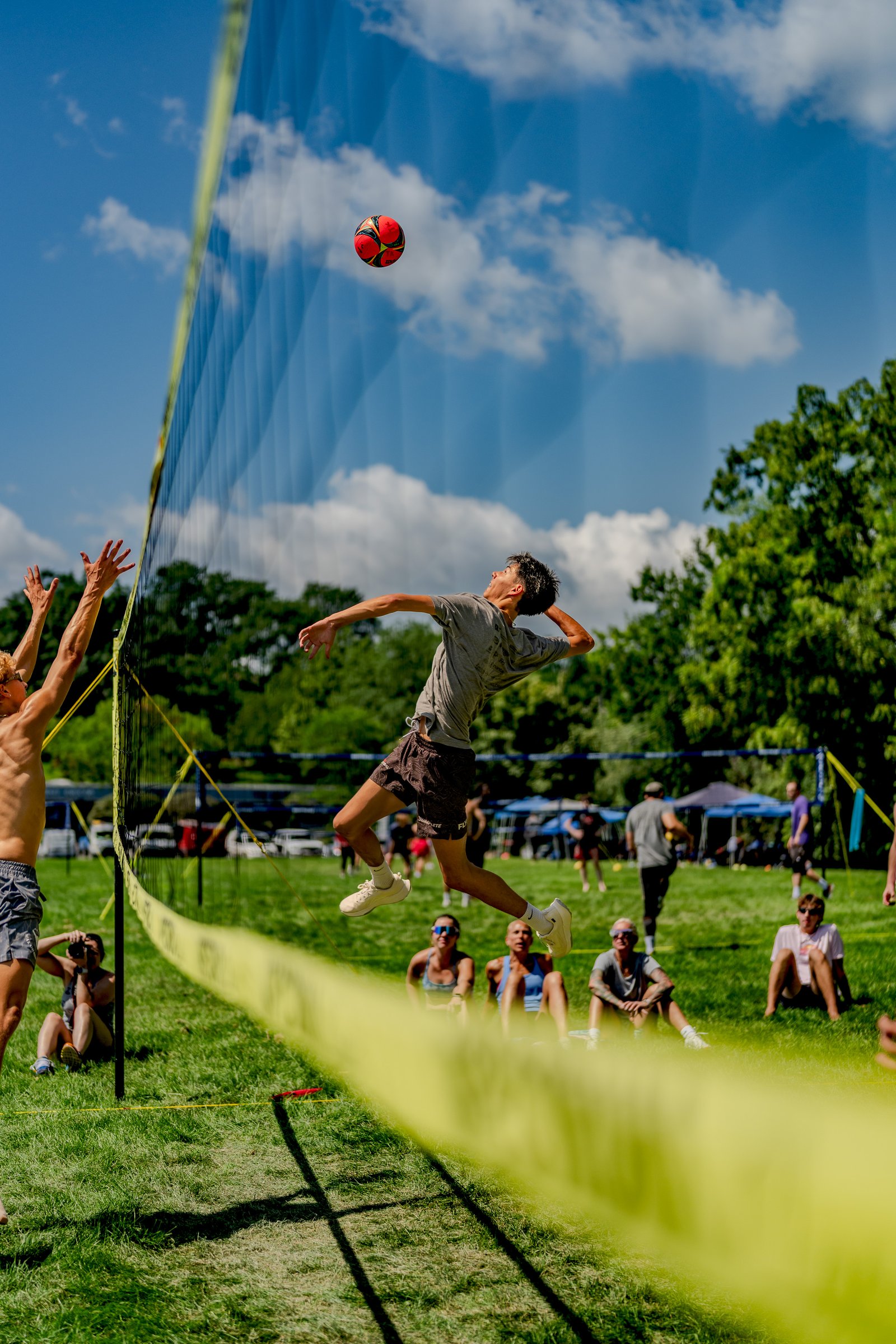 A player in a grey shirt spikes a volleyball over the net while another player in a grey shirt reaches up, with spectators watching from the grass.