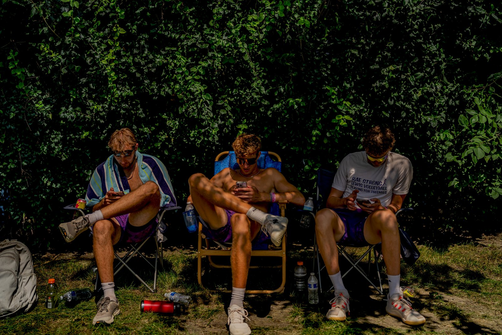 Three players in purple shorts sit in chairs, looking at their phones, with a leafy green background.