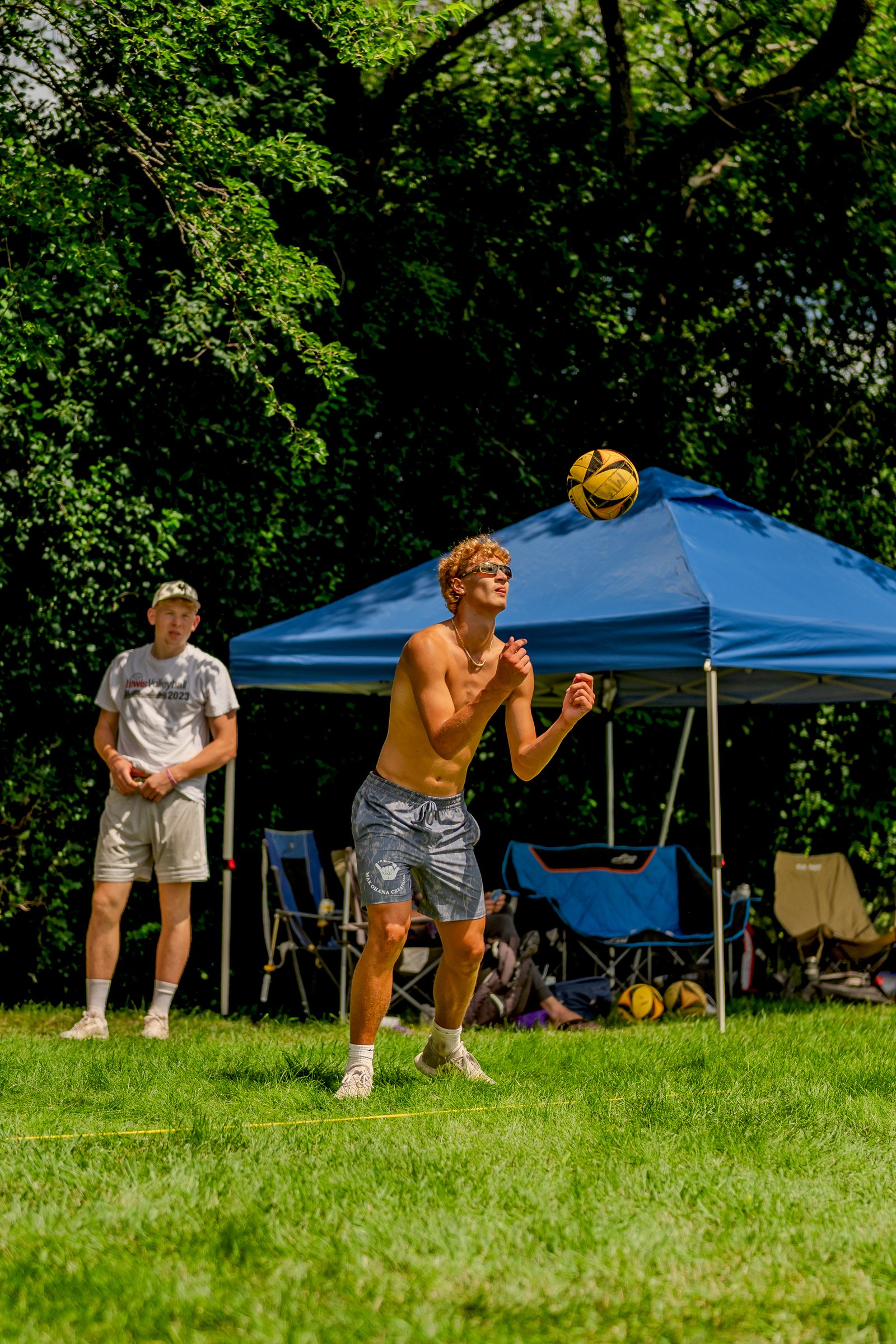 A player in grey shorts prepares to hit a volleyball as another player in a white shirt watches, with a blue tent in the background.