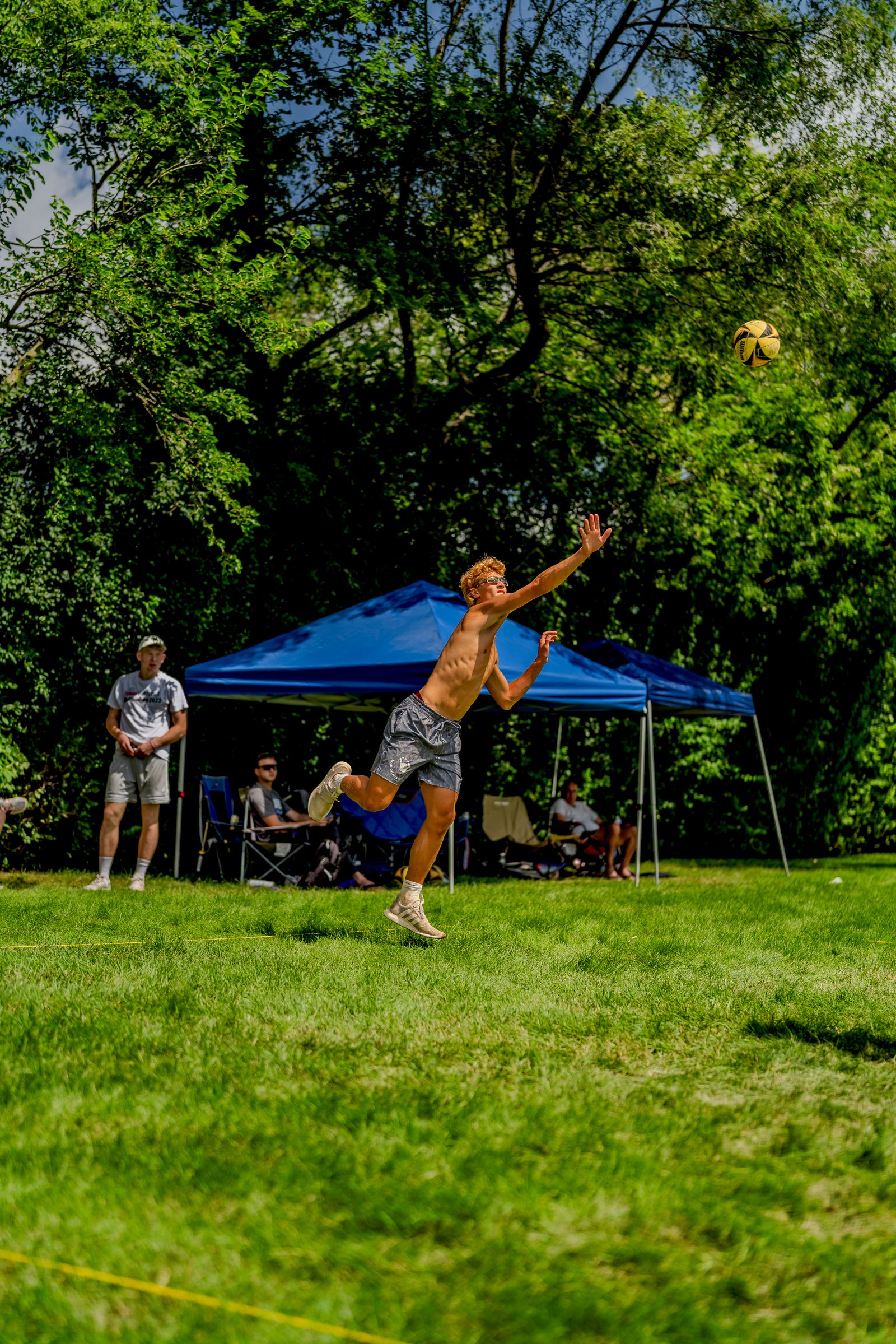 A player in grey shorts jumps to hit a volleyball during a tournament, with spectators under a blue tent.
