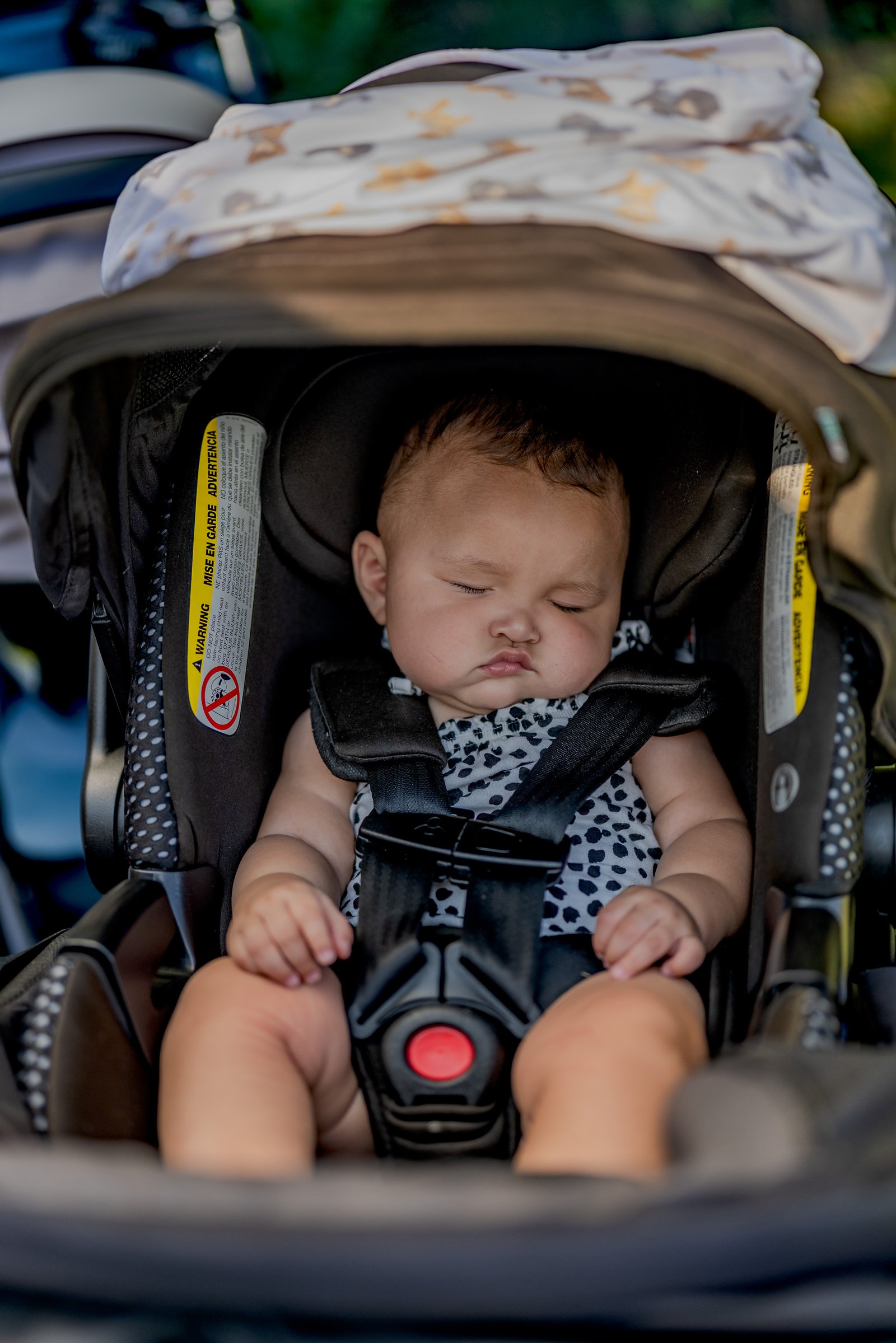 A baby sleeps in a car seat with a black and white spotted outfit and a patterned blanket.