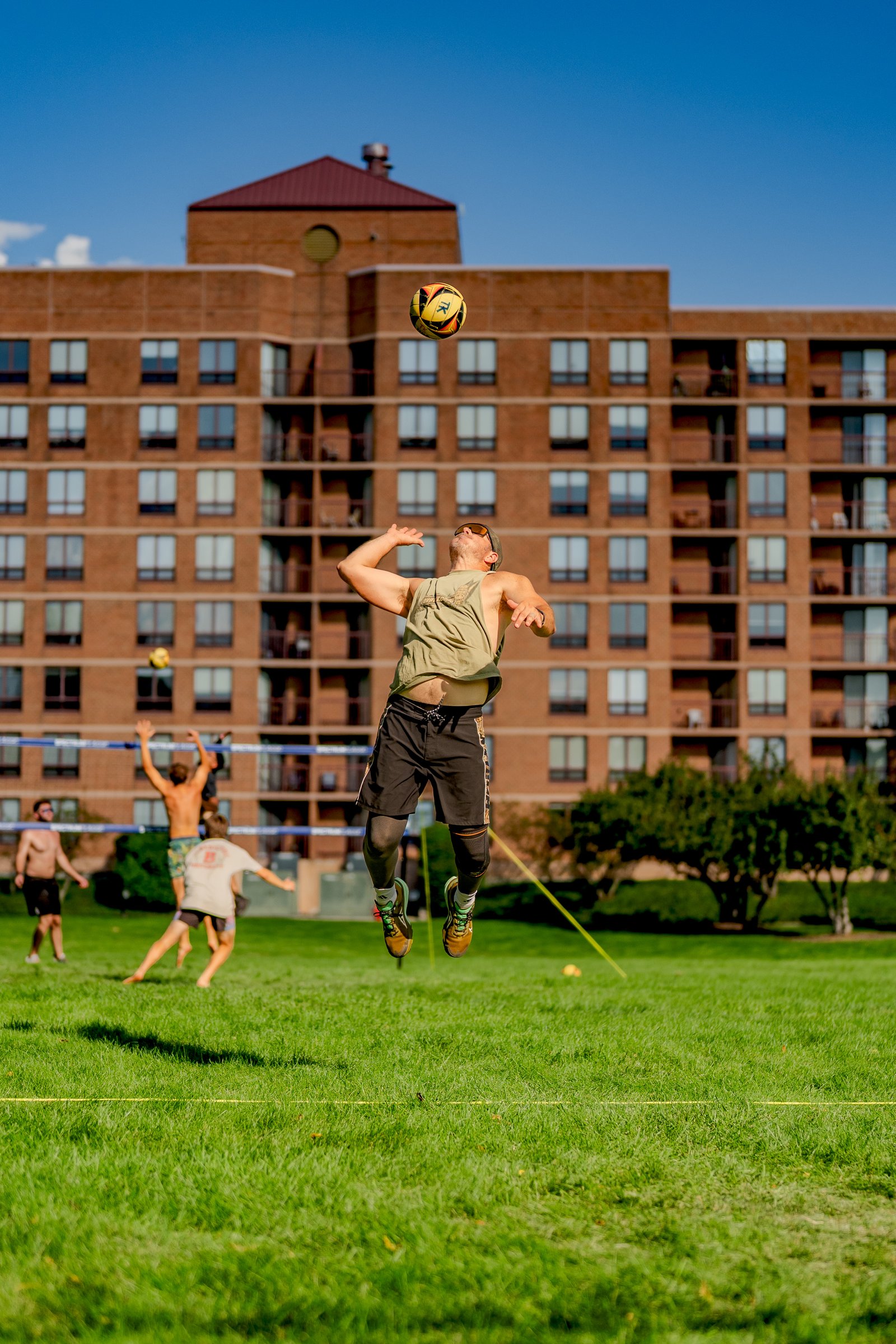 A player in a green tank top jumps to hit a volleyball over the net, with other players in the background on a grassy court.