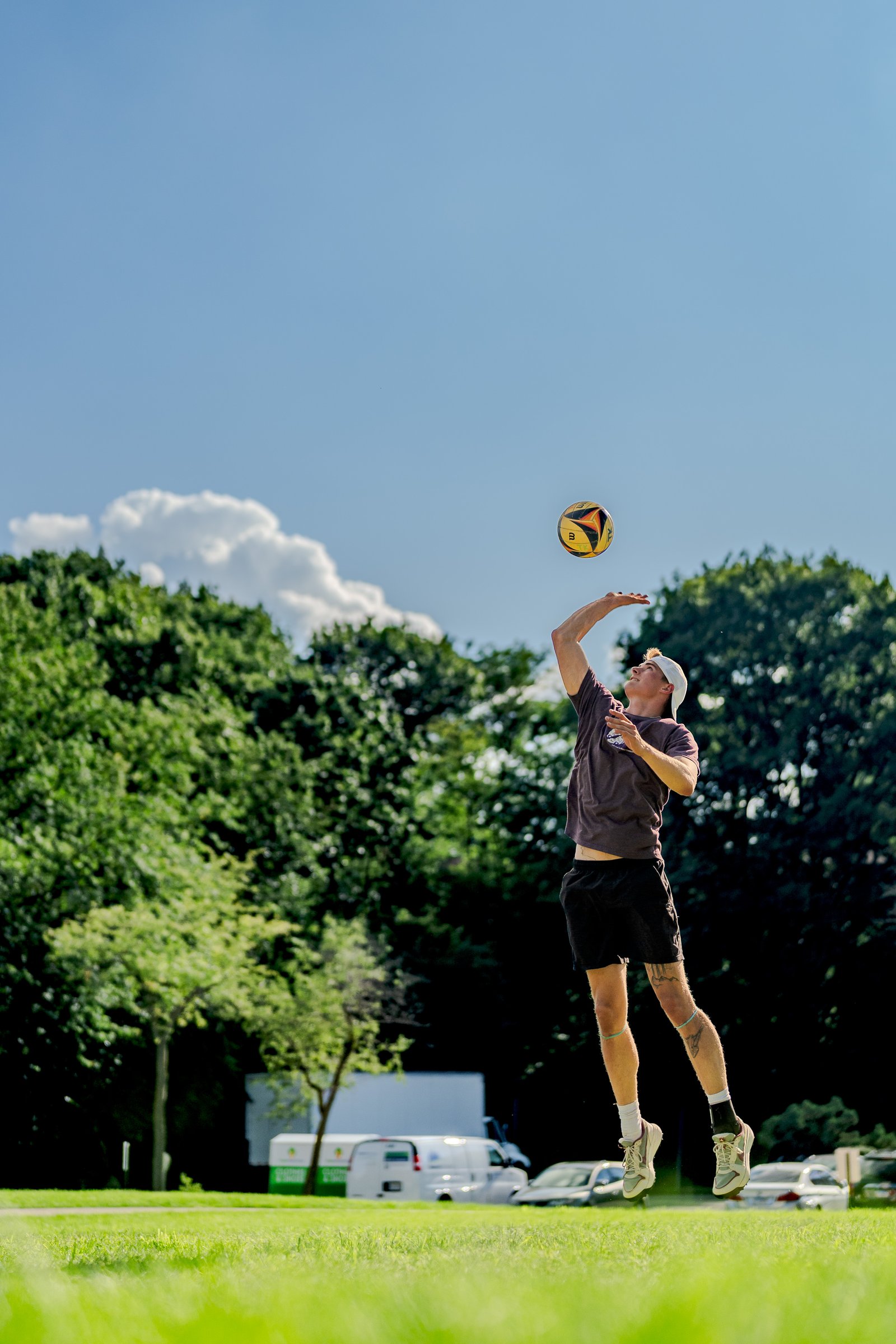 A player in a dark shirt jumps to hit a volleyball against a blue sky with trees in the background.