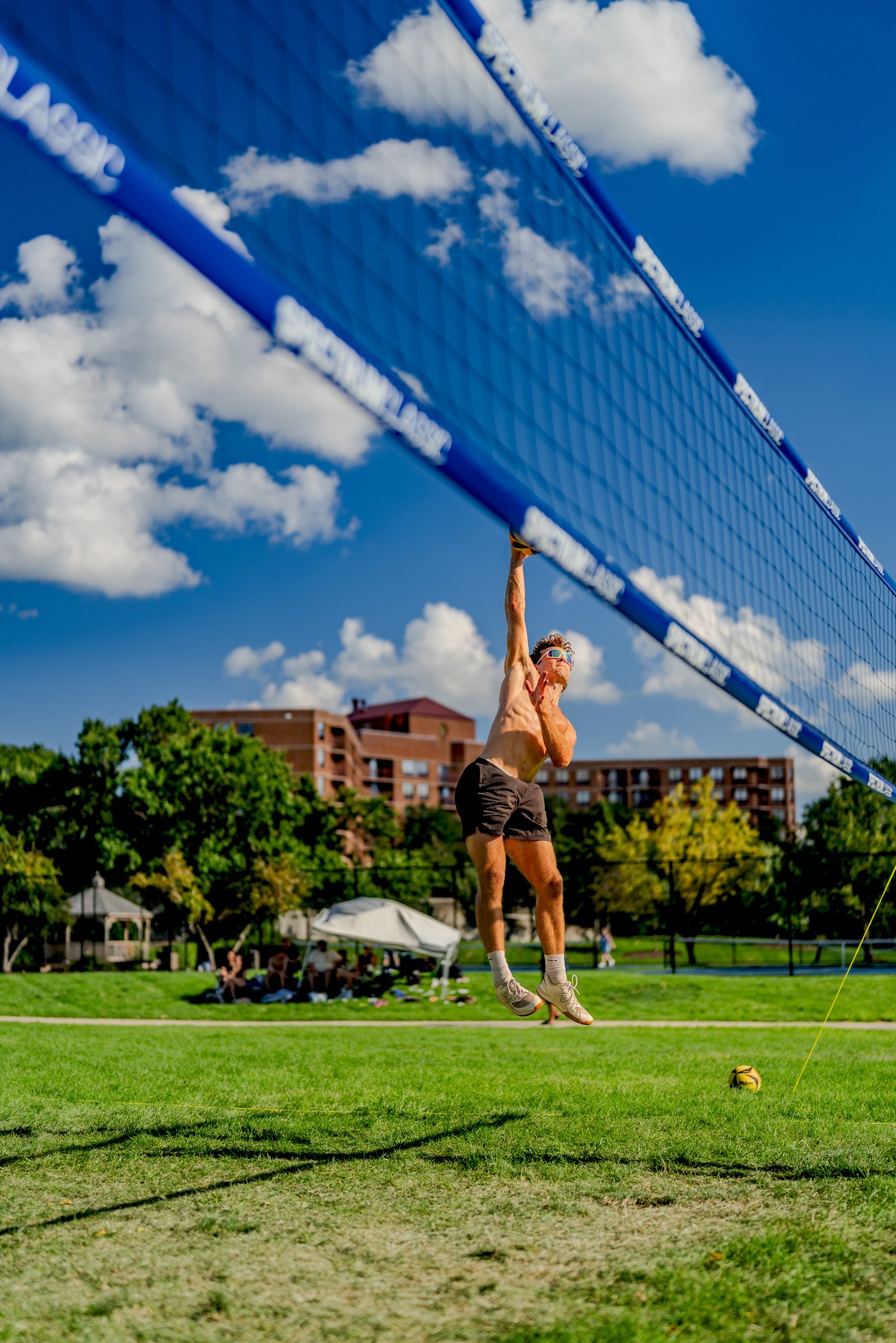 A player in black shorts jumps to hit a volleyball over the net, with spectators in the background.