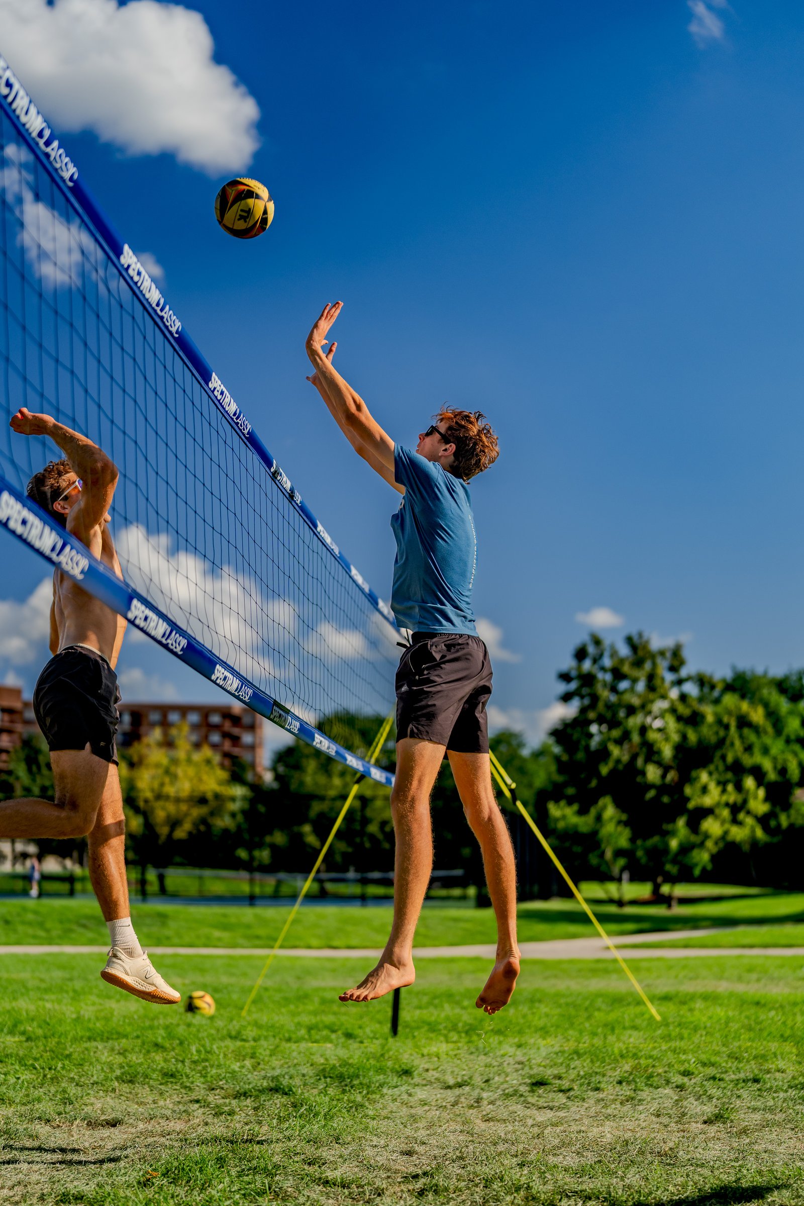 Two players jump near the net as a volleyball flies overhead on a sunny day.