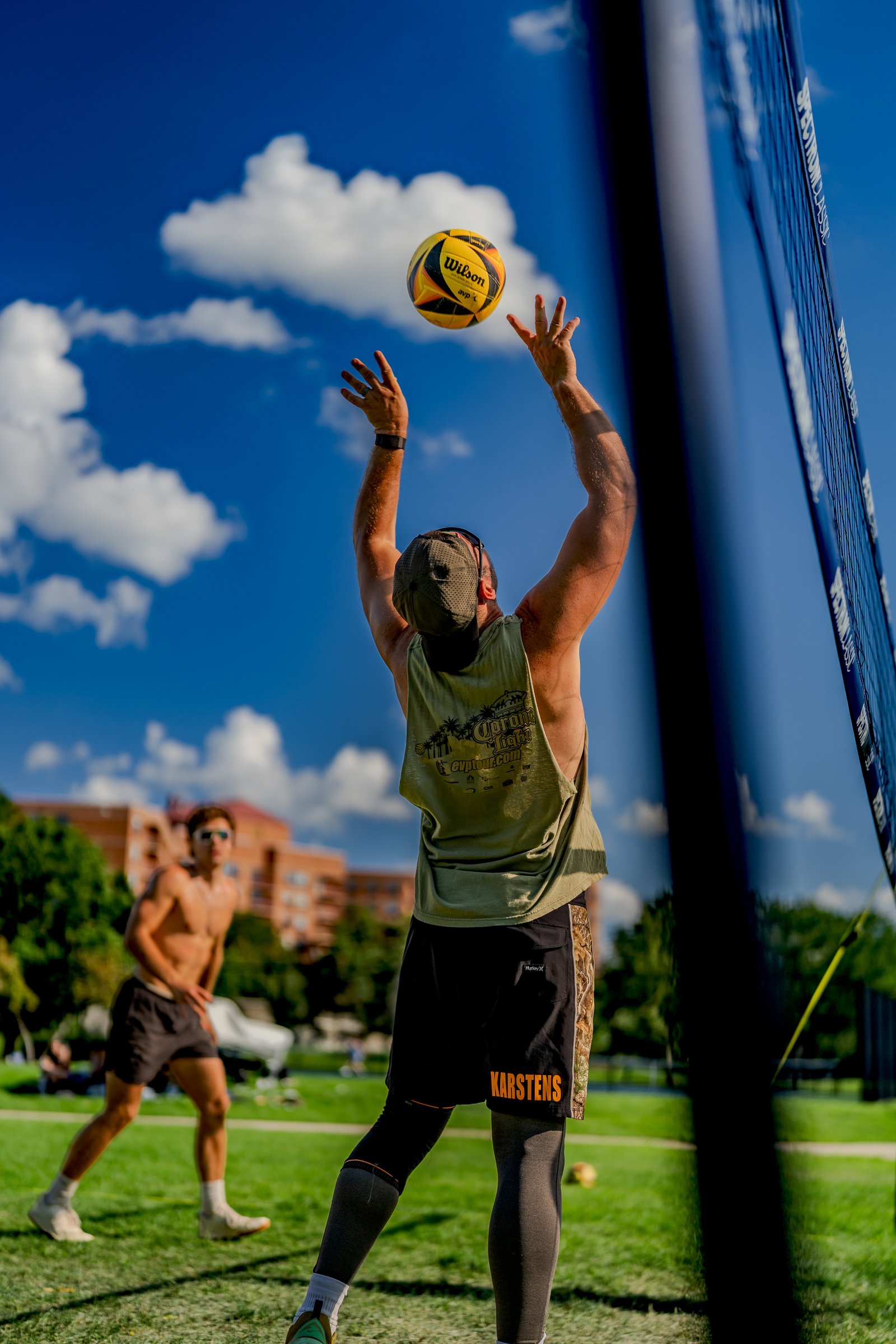 A player in a green tank top and black shorts with orange lettering prepares to hit a volleyball against a blue sky with clouds.