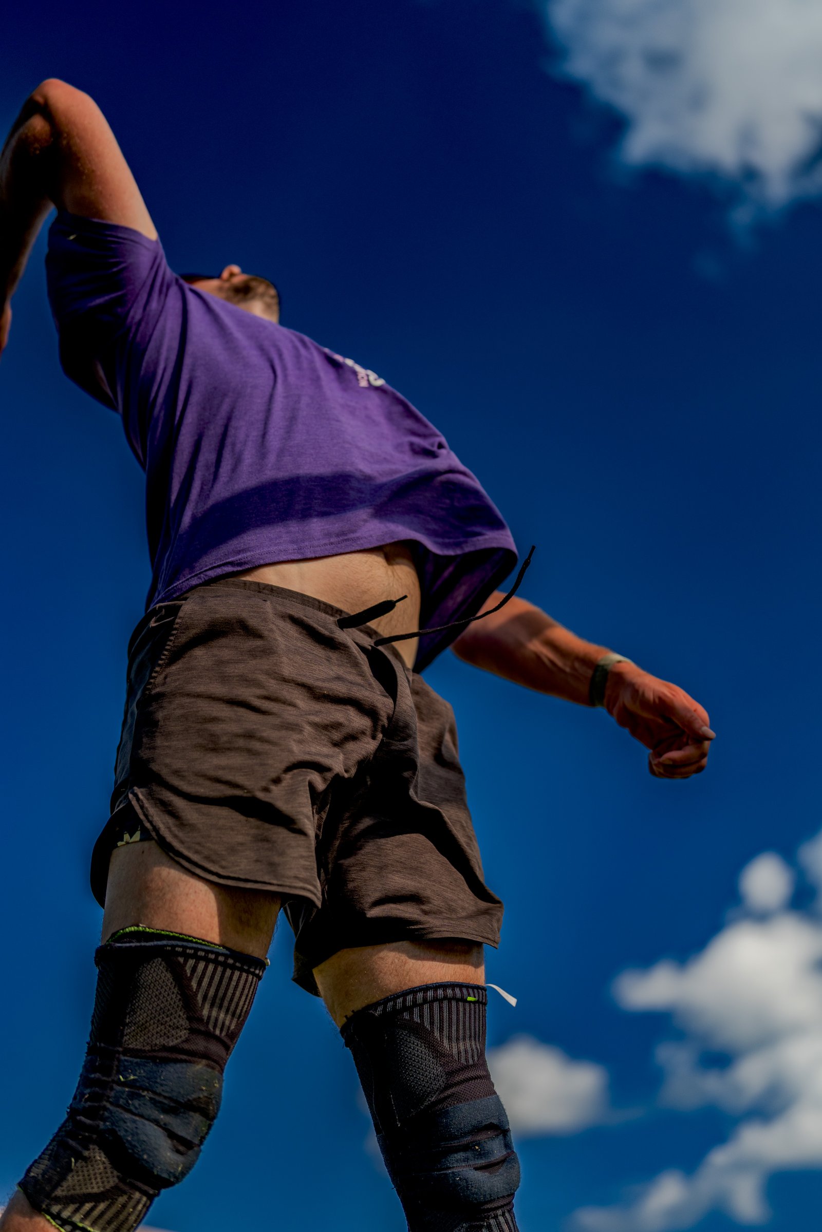 A player in a purple shirt and brown shorts jumps against a blue sky with clouds.