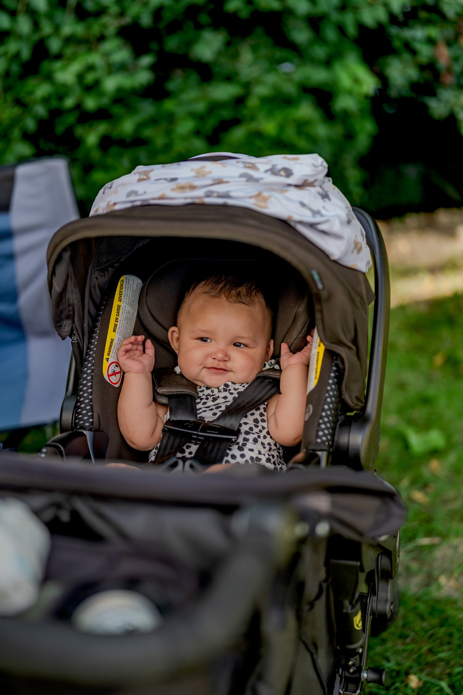A baby sits in a stroller with a patterned blanket and a warning label.