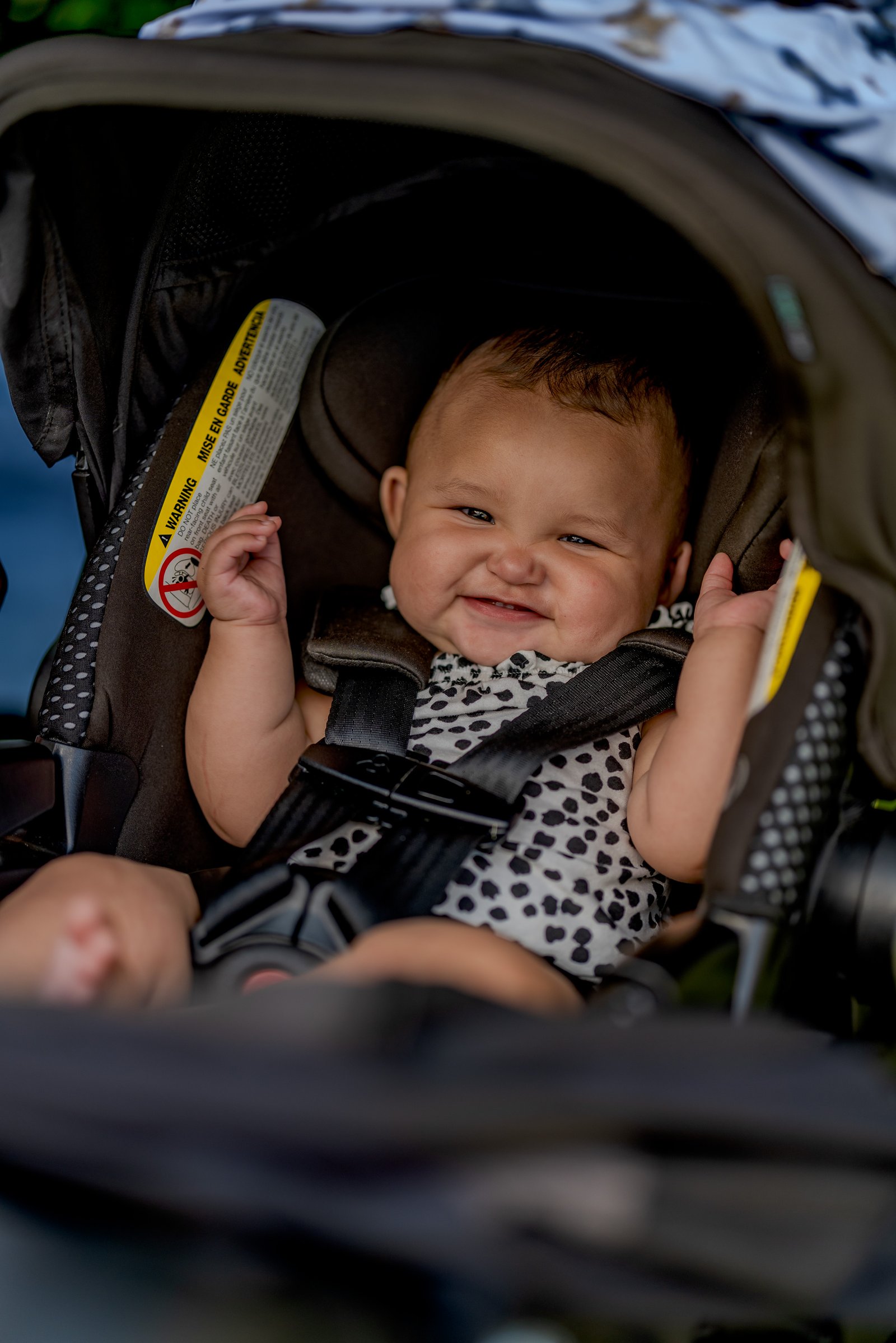 A baby smiles in a stroller with a warning label visible.