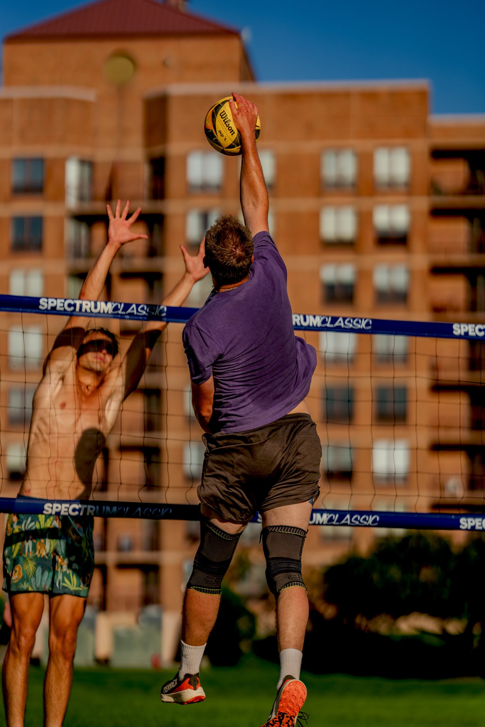 A player in a purple shirt jumps to hit a volleyball over the net, with another player in patterned shorts reaching up to block.