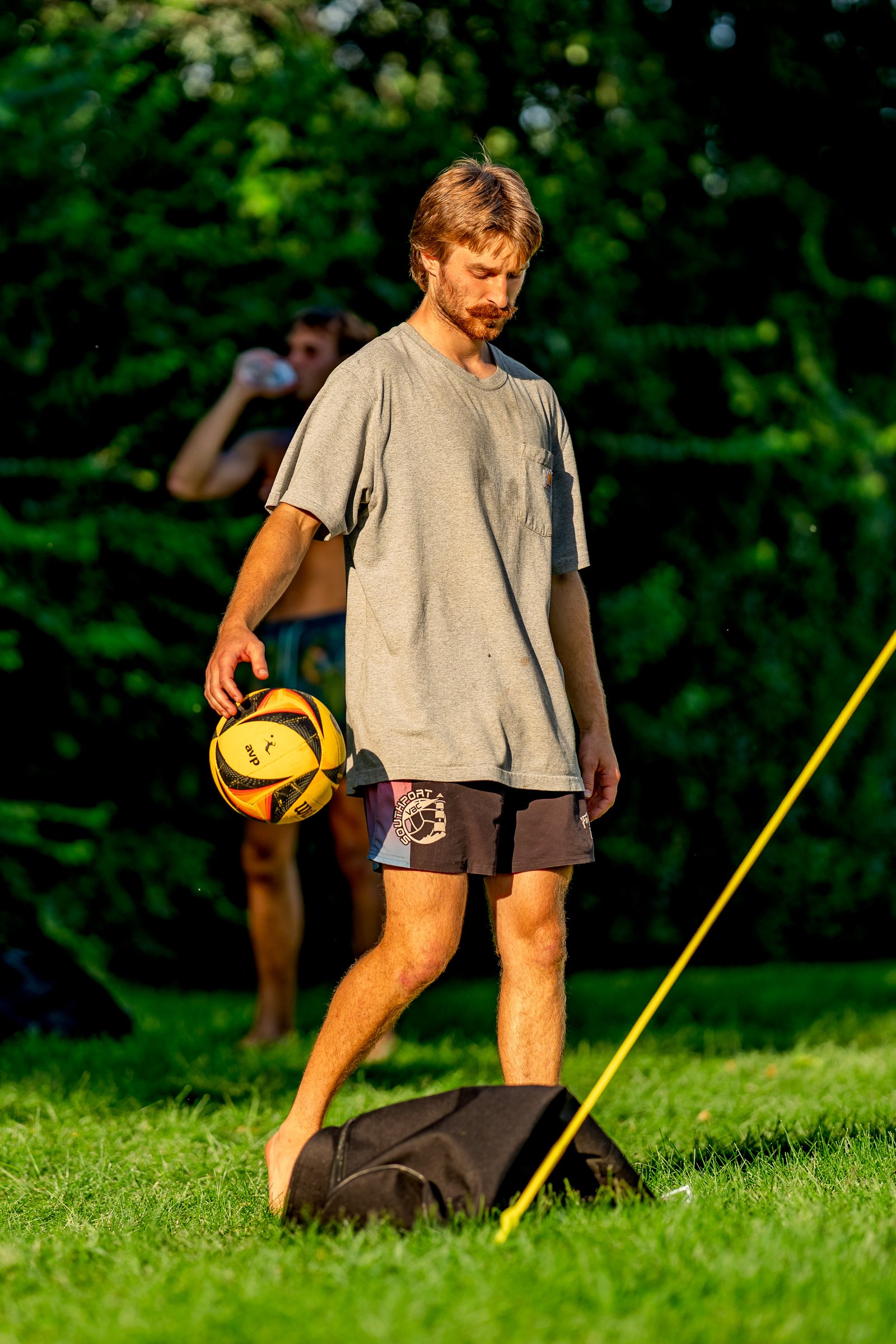A player in a grey shirt and dark shorts holds a volleyball while another player drinks in the background at a tournament.